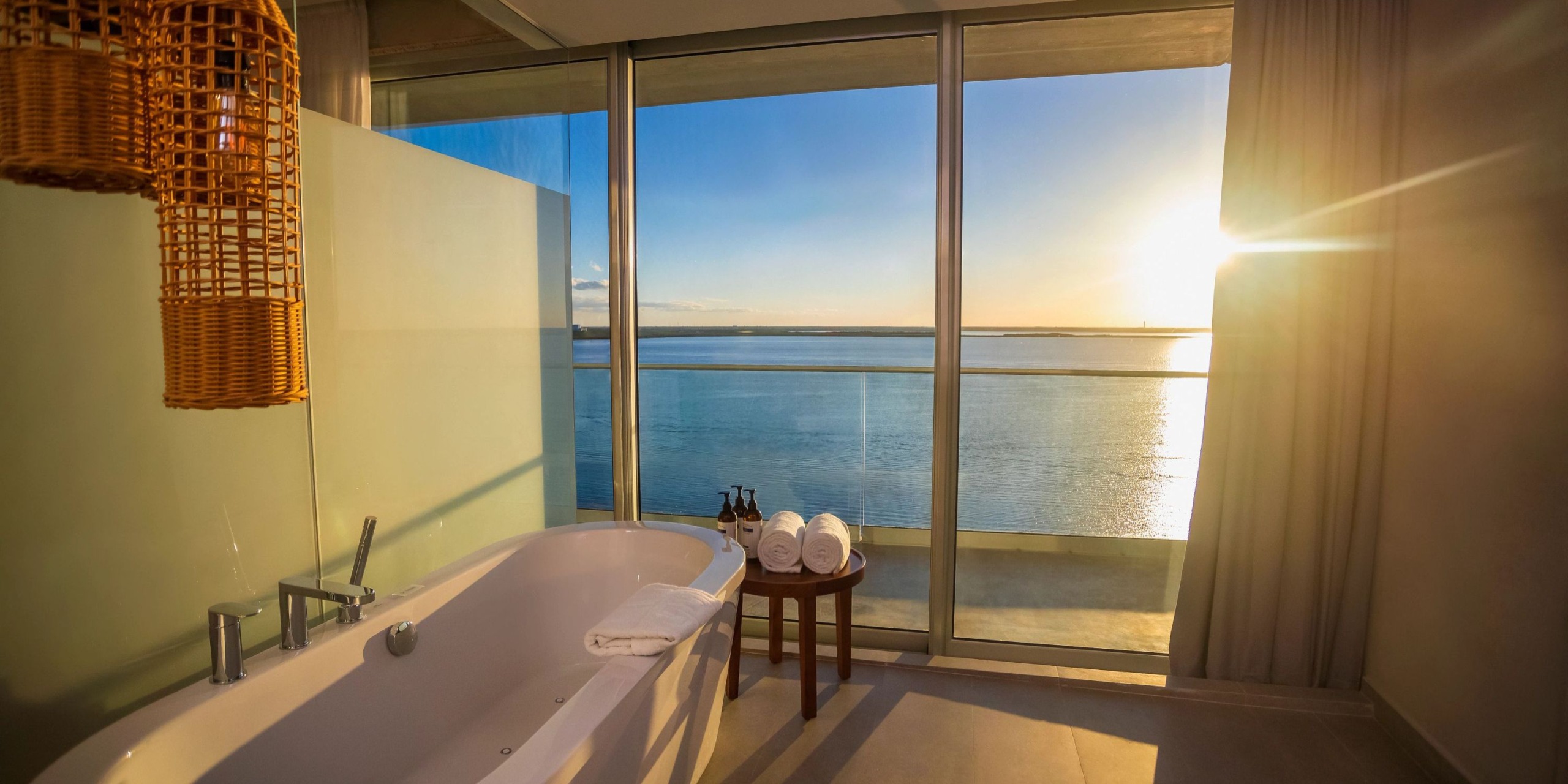 A bathroom in the Hyatt Vivid Grand Island features a white bathtub, rolled towels on a small table, and offers a stunning view of the ocean through large windows at sunset.