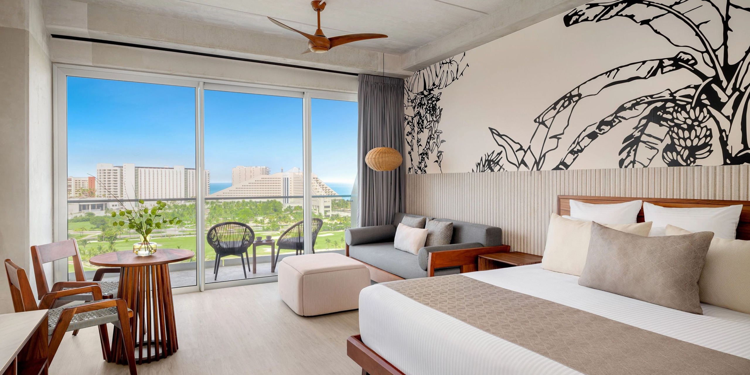 Experience the Hyatt Vivid Grand Island: a hotel room featuring a king-sized bed, a comfortable sofa, and a small table with chairs. Enjoy breathtaking cityscape and ocean views through the large window. The room’s wall features an elegant botanical design, complemented by modern decor in neutral tones.