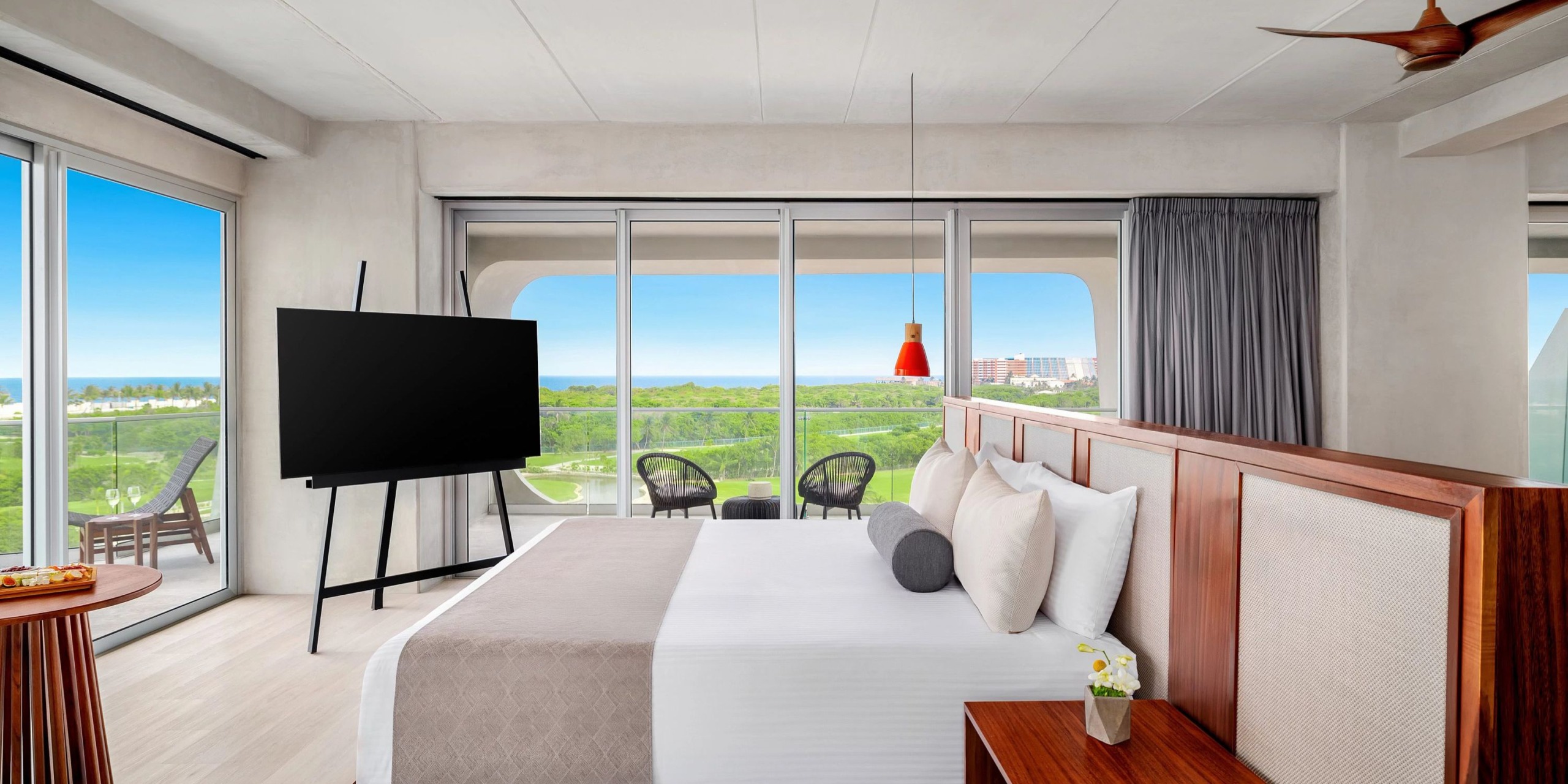 Experience the elegance of the Hyatt Vivid Grand Island, featuring a contemporary hotel room with a spacious bed, a flat-screen TV, and expansive floor-to-ceiling windows that provide access to a balcony outfitted with outdoor chairs and lush greenery, offering a scenic view of distant city buildings.
