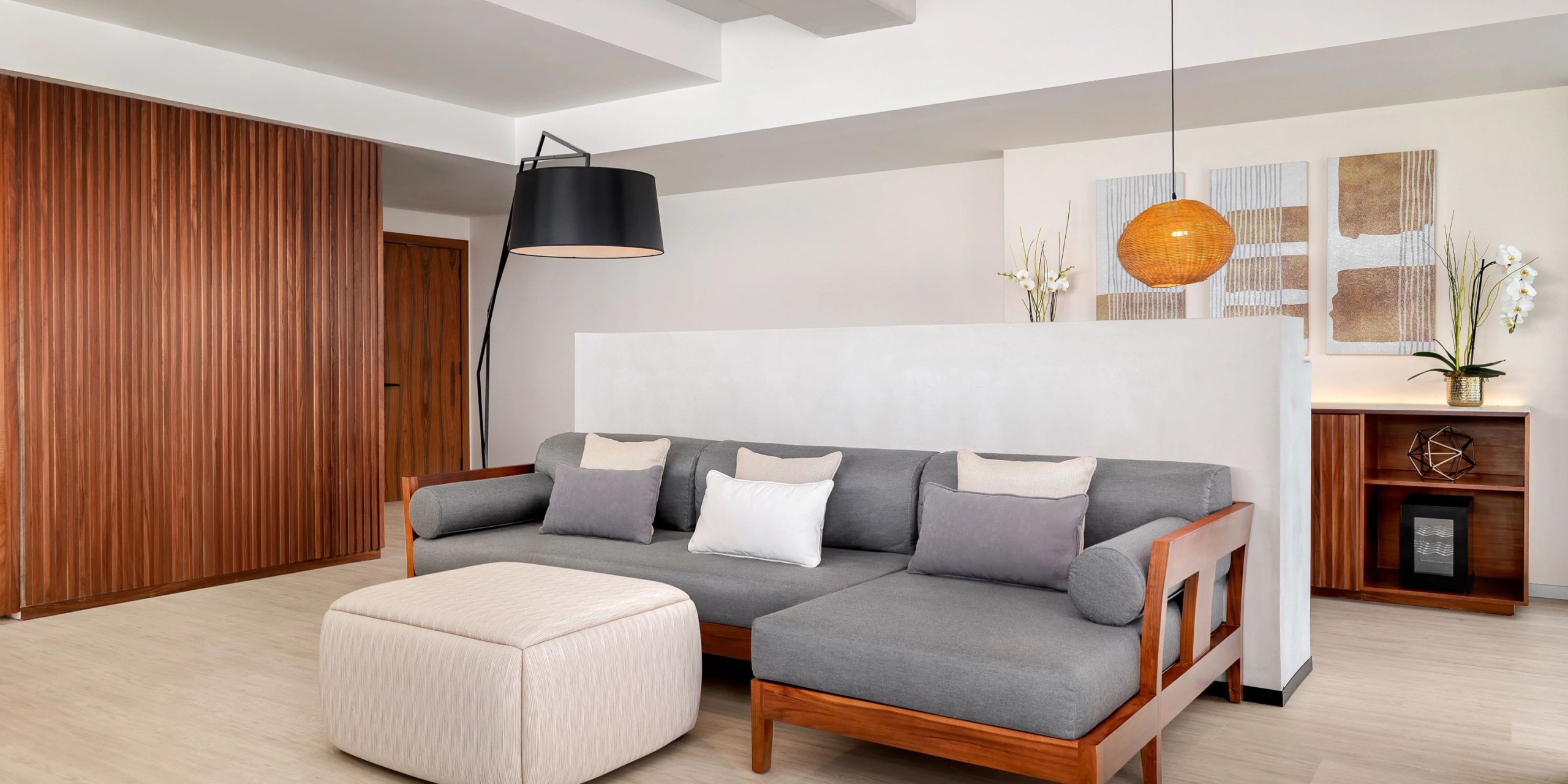 A modern living room featuring the Hyatt Vivid Grand Island sectional sofa in gray, paired with a beige ottoman, complemented by a floor lamp and pendant light. Wooden accents and minimalist wall art beautifully complete the decor.