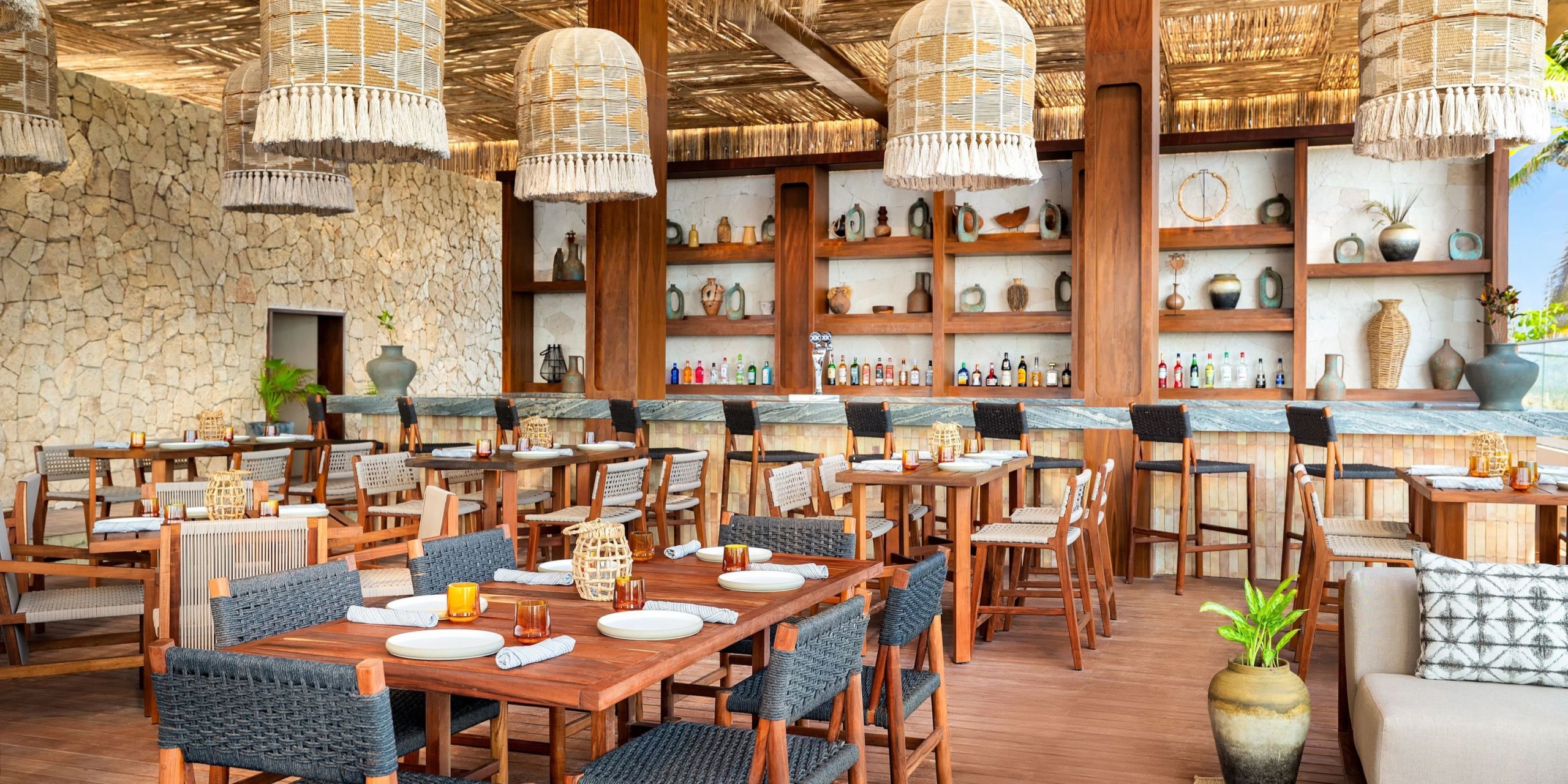 The Hyatt Vivid Grand Island features an outdoor dining area with wooden tables and chairs, complemented by wicker light fixtures. Its bar boasts a selection of various bottles displayed on shelves, while the decor includes pottery and lush plants.