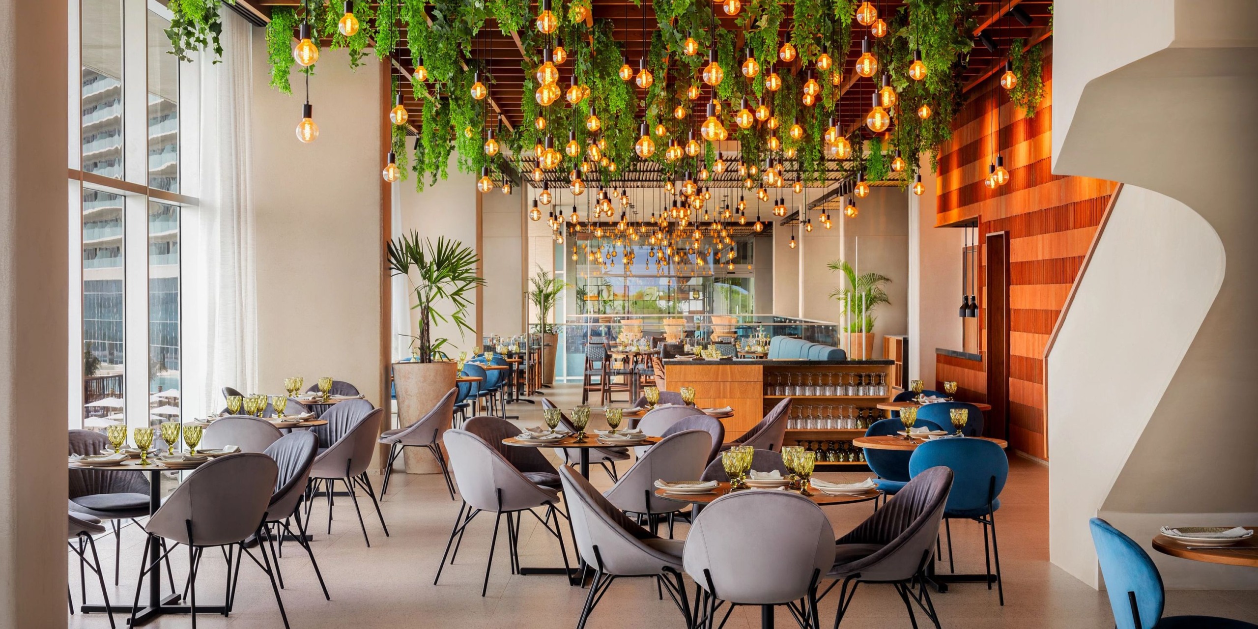 The Hyatt Vivid Grand Island exudes a contemporary ambiance with its gray chairs, wooden accents, and suspended greenery. Round tables are elegantly set with tableware, while warm globe lights dangle from the ceiling. Expansive windows allow for an abundance of natural light to pour in.