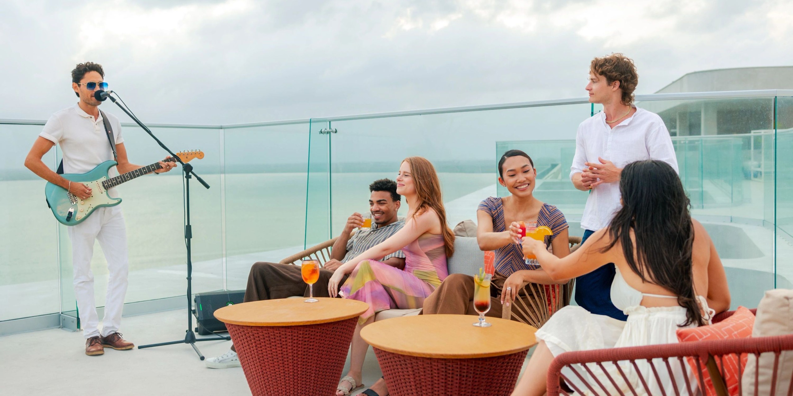 A group of people enjoy drinks on the rooftop of Hyatt Vivid Grand Island. A musician plays guitar while others sit and chat around the outdoor tables, with a cloudy sky in the background.