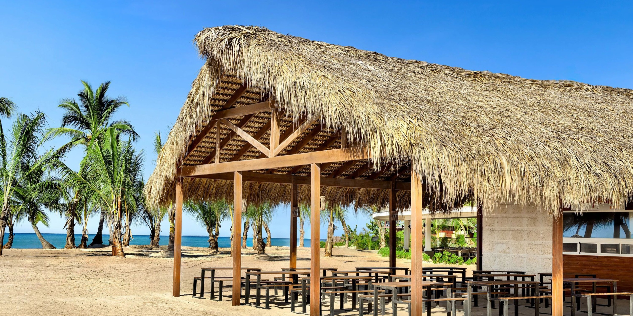 The Sunscape Dominicus La Romana features a thatched-roof beach pavilion with wooden beams and seating, nestled among palm trees near the ocean under a clear blue sky.