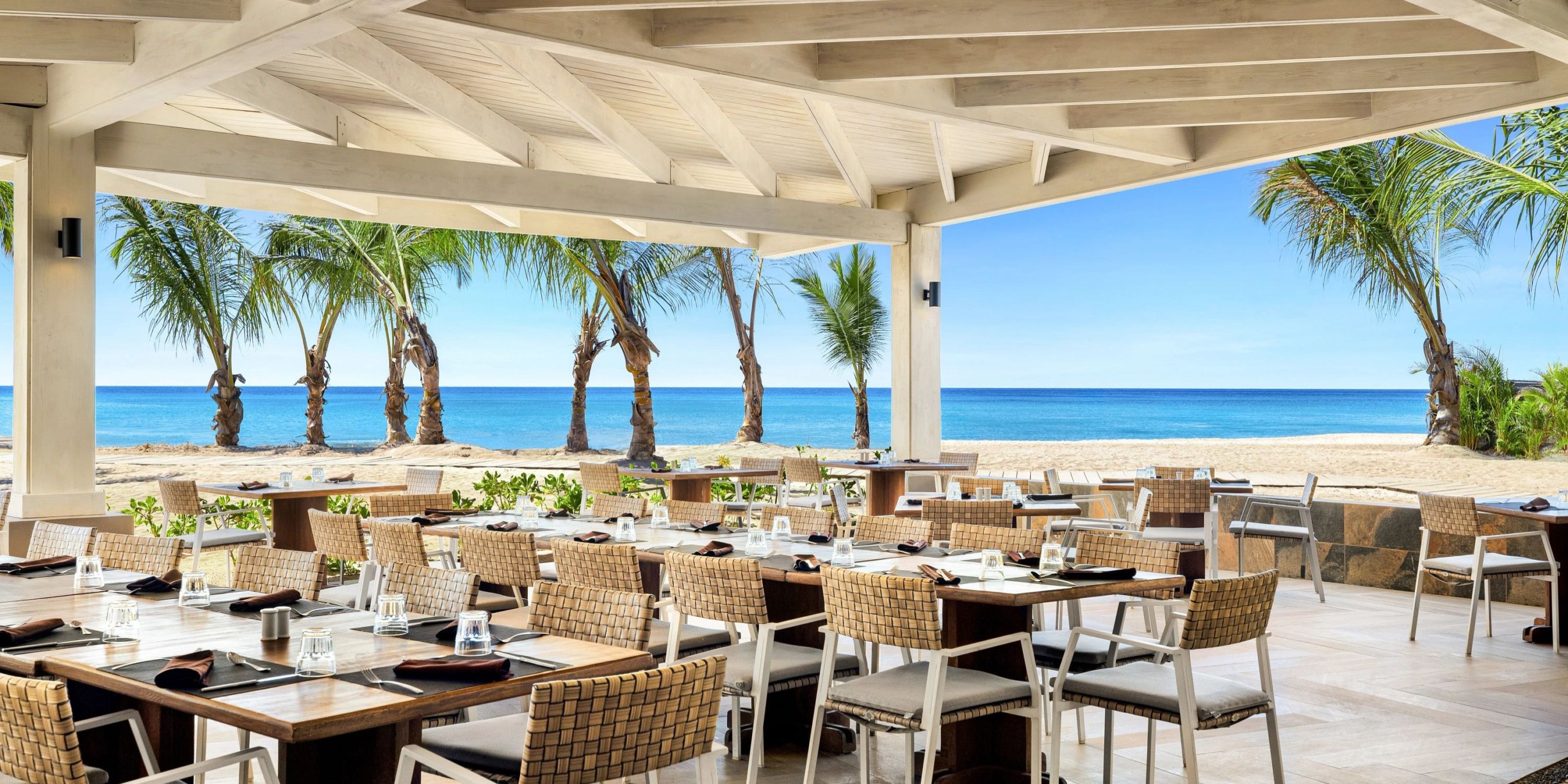 Enjoy dining at the Sunscape Dominicus La Romana's beachside restaurant, featuring tables and chairs nestled under a wooden pergola with stunning views of palm trees and the ocean.