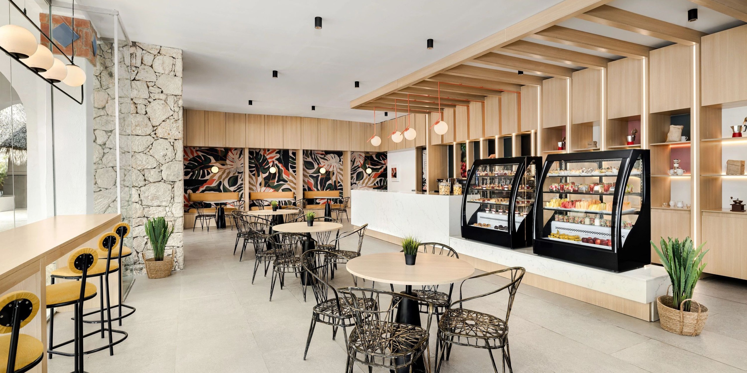 The Sunscape Dominicus La Romana features a contemporary cafe design with round tables and chairs, a stone-accented wall, display cases filled with snacks, and a ceiling crafted from wood. Pendant lights gracefully suspend from above, infusing the space with a welcoming warmth.