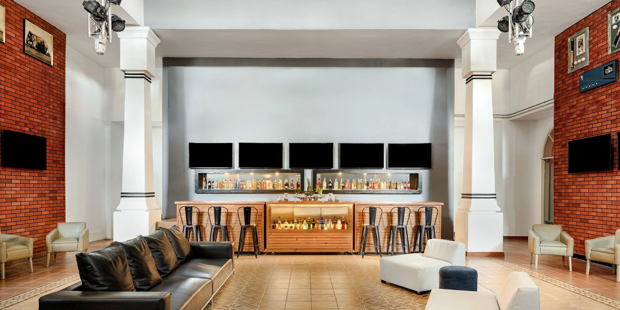 Experience the contemporary bar interior at Sunscape Dominicus La Romana, featuring a central bar counter, high stools, a comfortable sofa, and framed pictures adorning the brick walls. Above the bar, four screens are strategically mounted for your viewing pleasure.