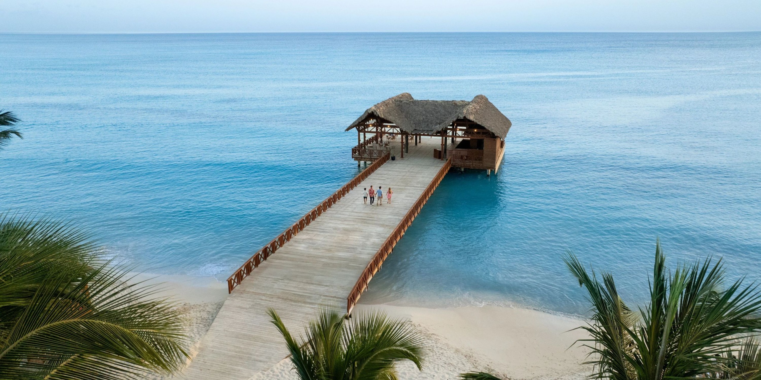 At Sunscape Dominicus La Romana, a wooden pier stretches over the azure waters leading to a charming thatched-roof hut, with a few visitors enjoying the view. Palm trees elegantly frame the scene in the foreground.