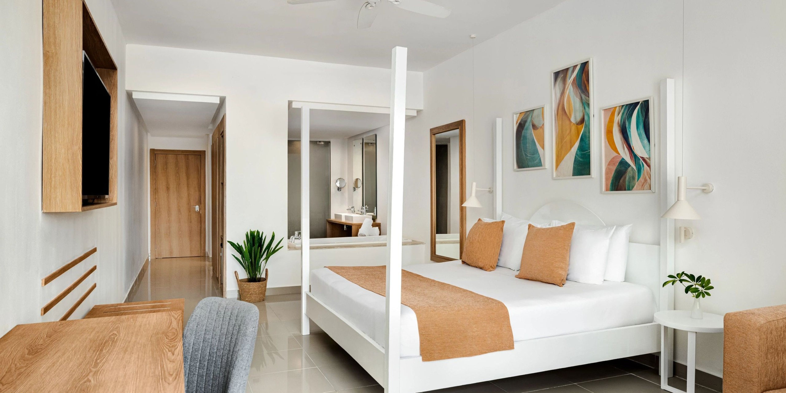 A luminous room at Sunscape Dominicus La Romana features a large bed adorned with orange accents, abstract art gracing the walls, and includes amenities like a desk, chair, and a plant. The bathroom area is visible in the background with a mirror.