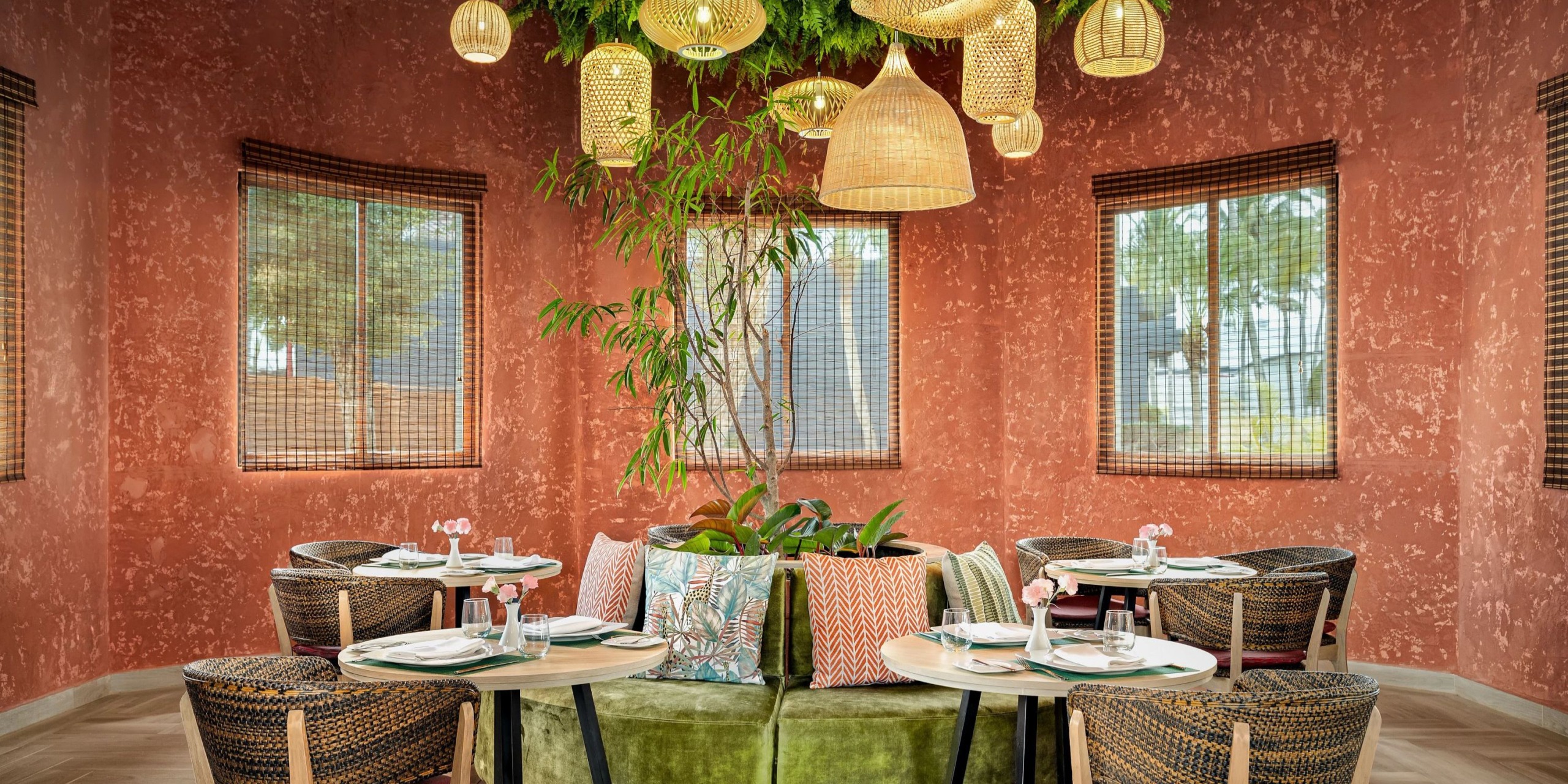 Experience a cozy dining atmosphere at Sunscape Dominicus La Romana, where round tables invite you for a delightful meal surrounded by stylish wicker chairs. At the heart of the setting, a central green booth showcases vibrant potted plants, while woven pendant lights cast a warm glow from above. The ambiance is enhanced by earth-toned walls and generous windows that contribute to the inviting charm.