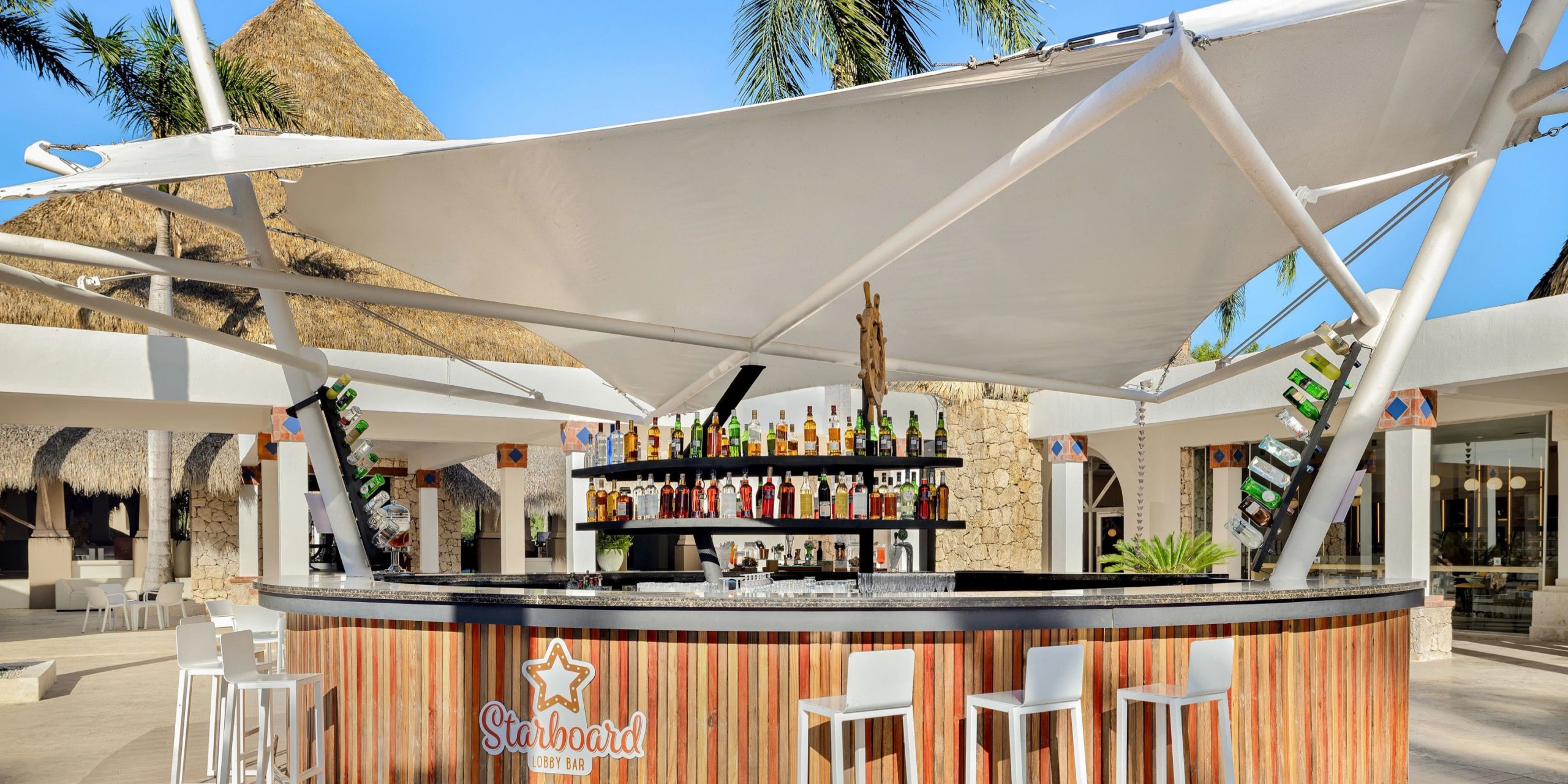 The Sunscape Dominicus La Romana features an outdoor bar with a wooden counter and white stools, complete with a canopy roof and shelves lined with various bottles, set against a backdrop of palm trees and tropical structures.