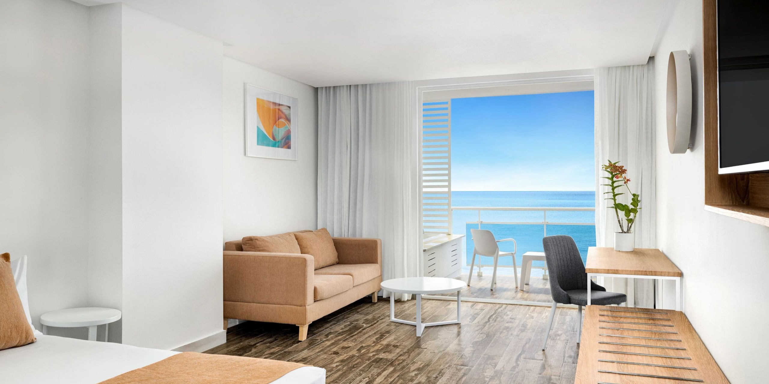 A contemporary room at Sunscape Dominicus La Romana includes a beige sofa, round table, desk, and chair. The space offers an expansive ocean view through a large window and comes equipped with a wall-mounted TV.