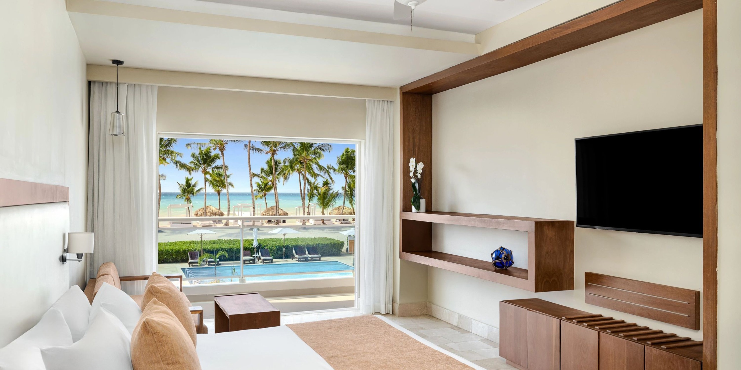 A contemporary room at Sunscape Dominicus La Romana offers a view of the pool and palm trees through its expansive window. The room is designed with white walls and includes a bed, TV, and sleek wooden shelves for a minimalist aesthetic.