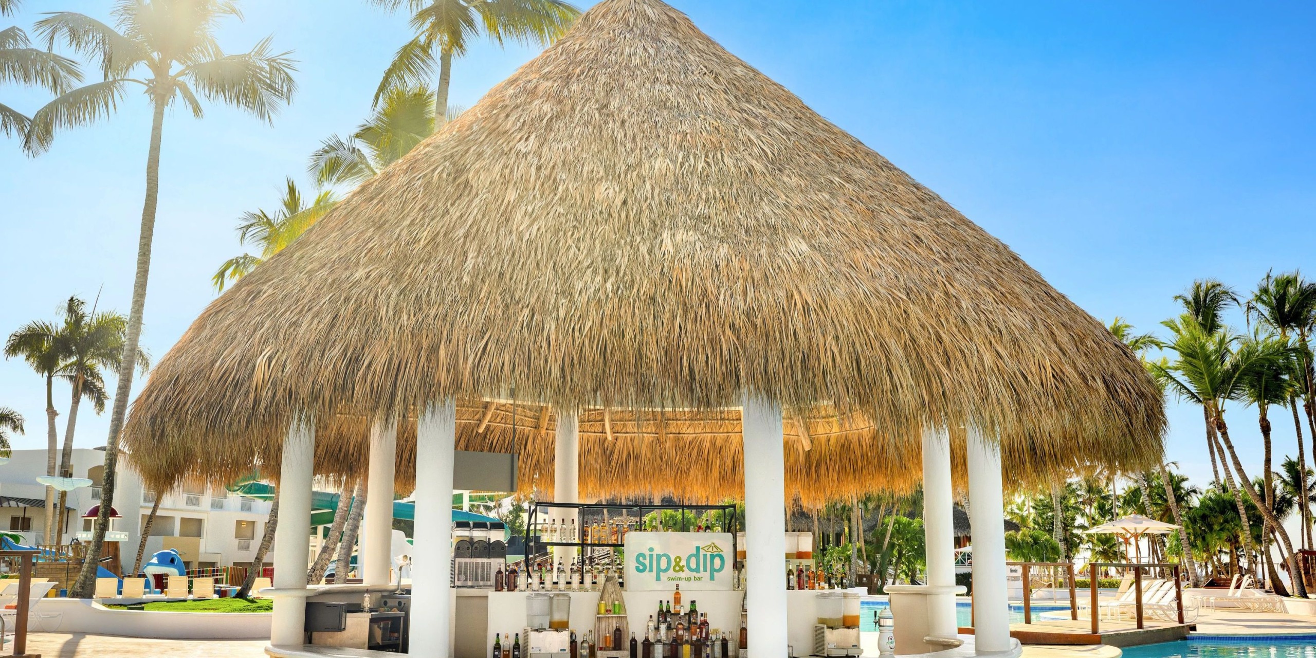 The Sunscape Dominicus La Romana features a tropical poolside bar with a thatched roof, encircled by palm trees, and provides an array of beverages beneath clear blue skies.