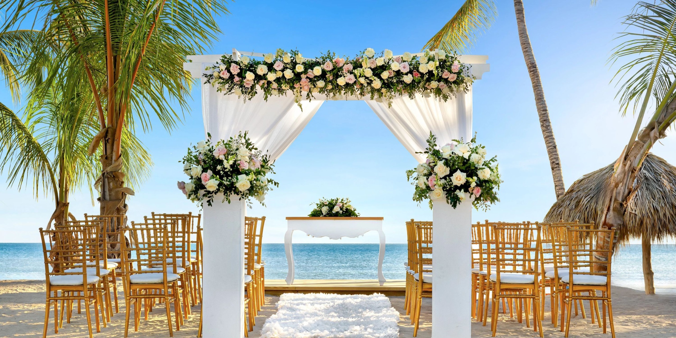 Sunscape Dominicus La Romana offers a beach wedding setup featuring a floral-decorated white arch, wooden chairs, and a small table set against the backdrop of clear blue skies and palm trees.