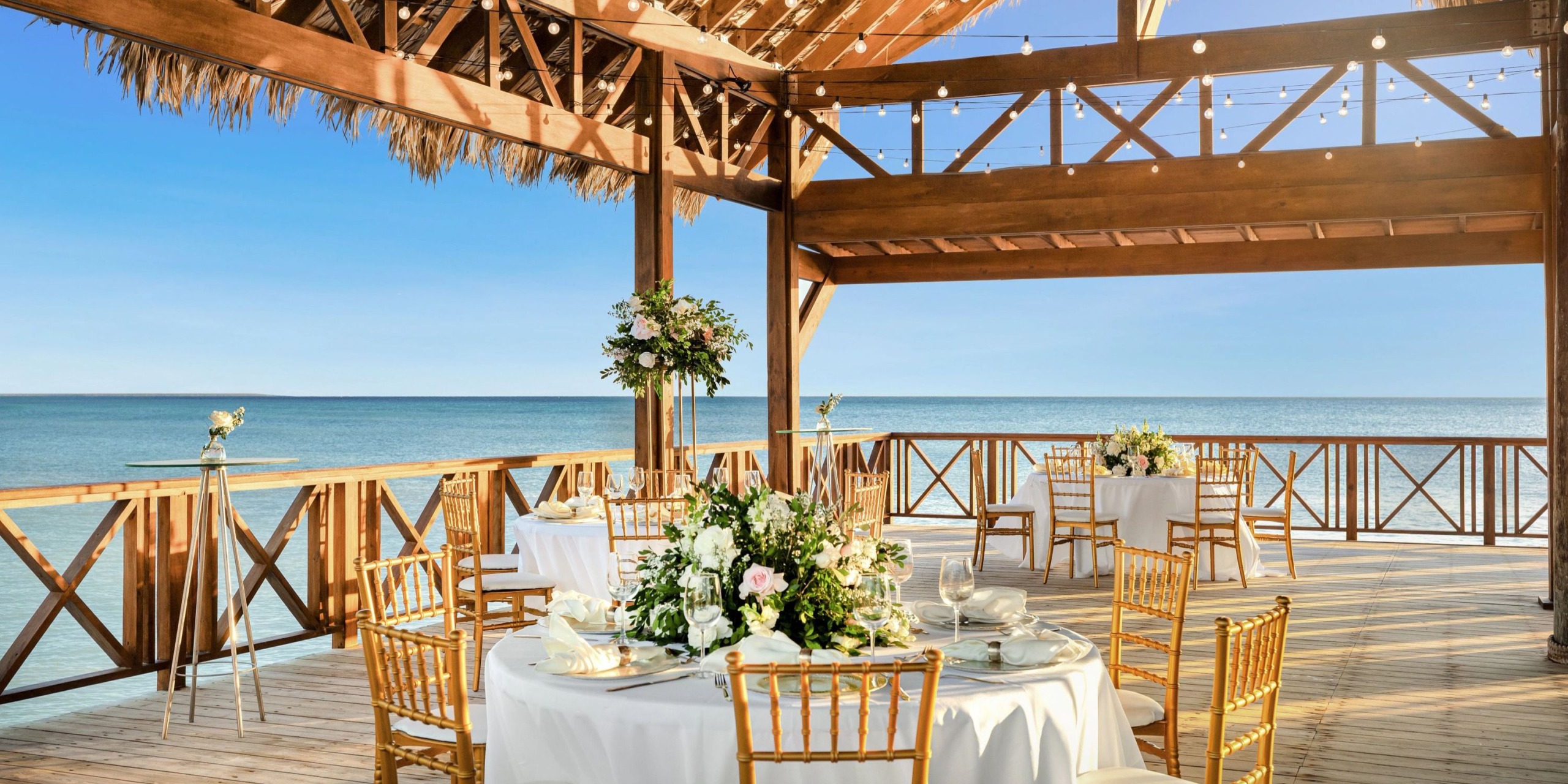 Enjoy an outdoor dining experience at Sunscape Dominicus La Romana, featuring round tables dressed in white tablecloths and adorned with floral centerpieces, all set beneath a charming wooden pergola with breathtaking views of the ocean.