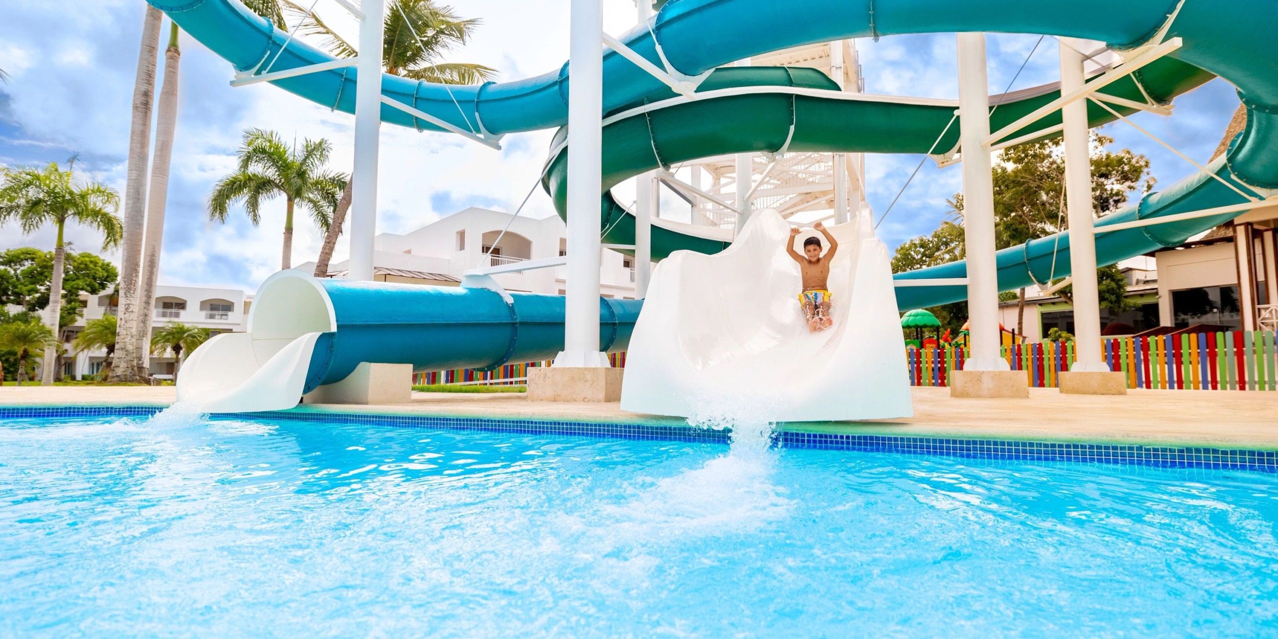 A person enjoying a ride down the vibrant, curvy green water slide at Sunscape Dominicus La Romana, splashing into the pool under the sunny sky, with picturesque palm trees and resort buildings in the background.