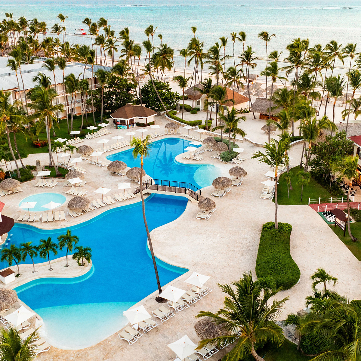Aerial view of Sunscape Coco Punta Cana, a tropical resort with blue swimming pools bordered by palm trees, sun loungers, and thatched umbrellas, all set against the backdrop of a beach and ocean.