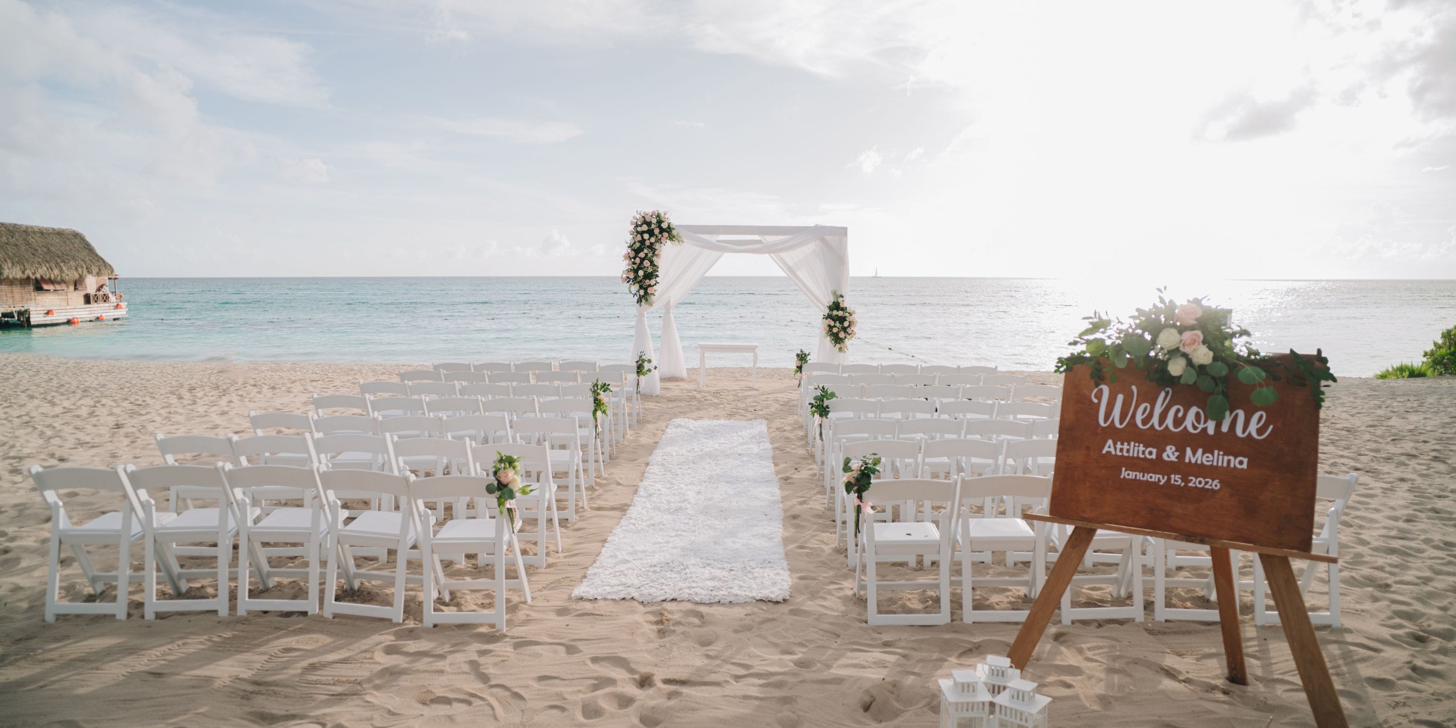 Destination wedding setup at Sunscape Dominicus La Romana: white chairs, floral arch, wood sign at sunset.