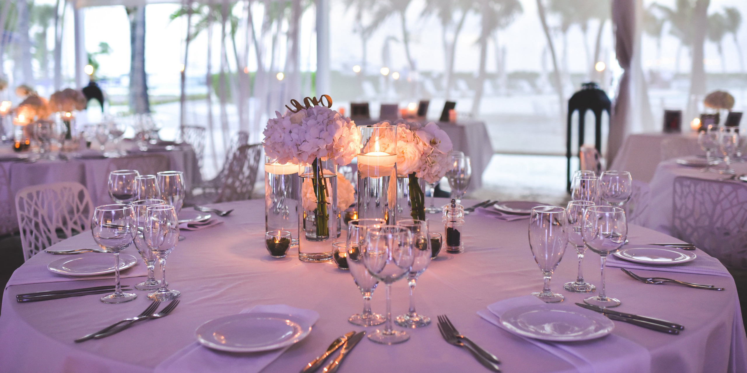 Experience an elegant table setting at the Alexandra Resort Turks and Caicos, featuring a canopy with white linens, glassware, and floral centerpieces, all nestled among trees in the gentle outdoor light.