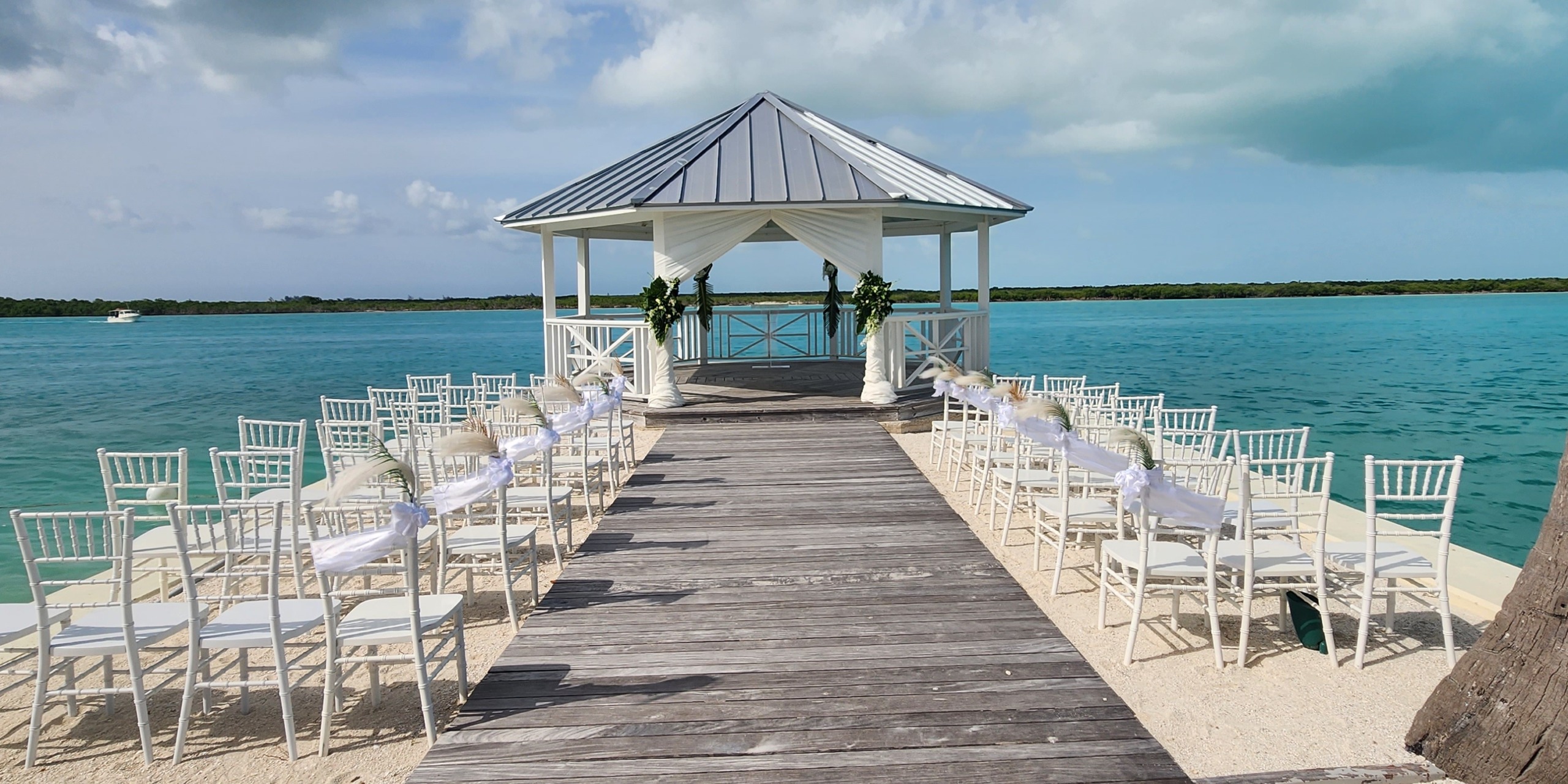 A wooden pier extends to a gazebo adorned for a wedding at the Alexandra Resort Turks and Caicos, surrounded by white chairs on a sandy beach with turquoise waters in the background.