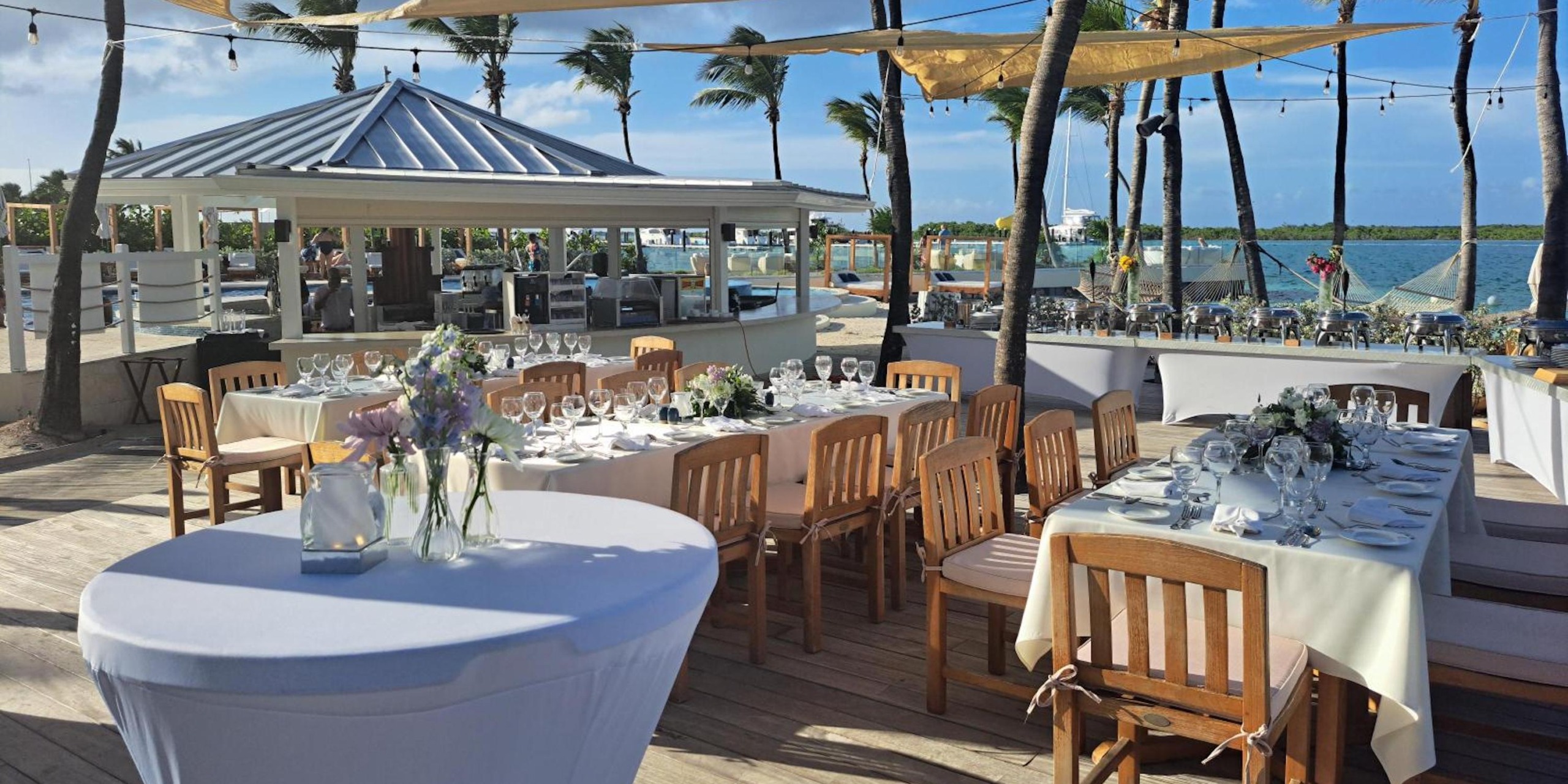 Enjoy an outdoor dining experience at the Alexandra Resort Turks and Caicos, complete with wooden tables and chairs under string lights and swaying palm trees by the beach.