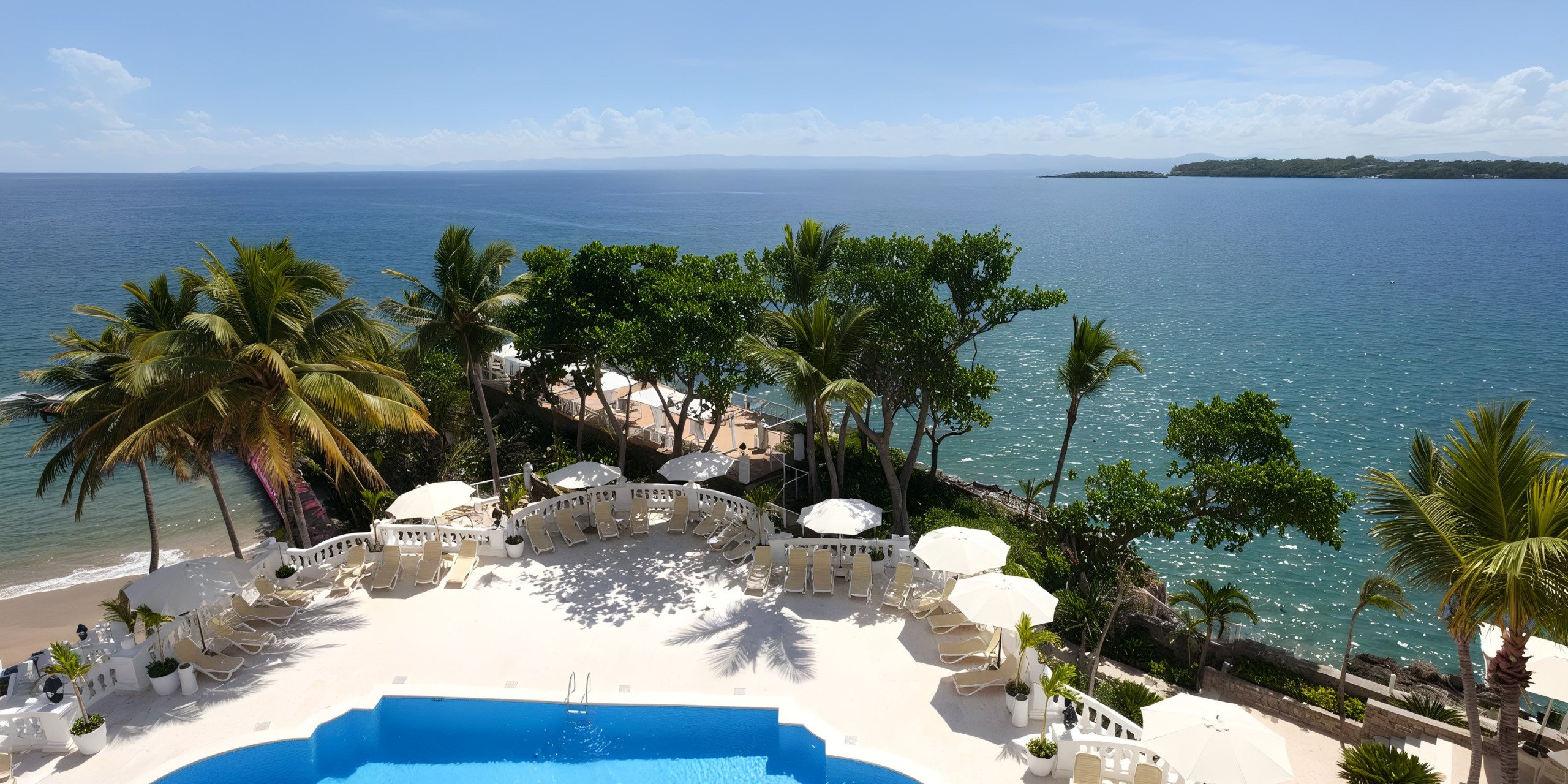 Aerial view of Bahia Principe Grand Samana, a coastal resort featuring a pool, palm trees, and lounge chairs that overlook a calm ocean under a clear blue sky.