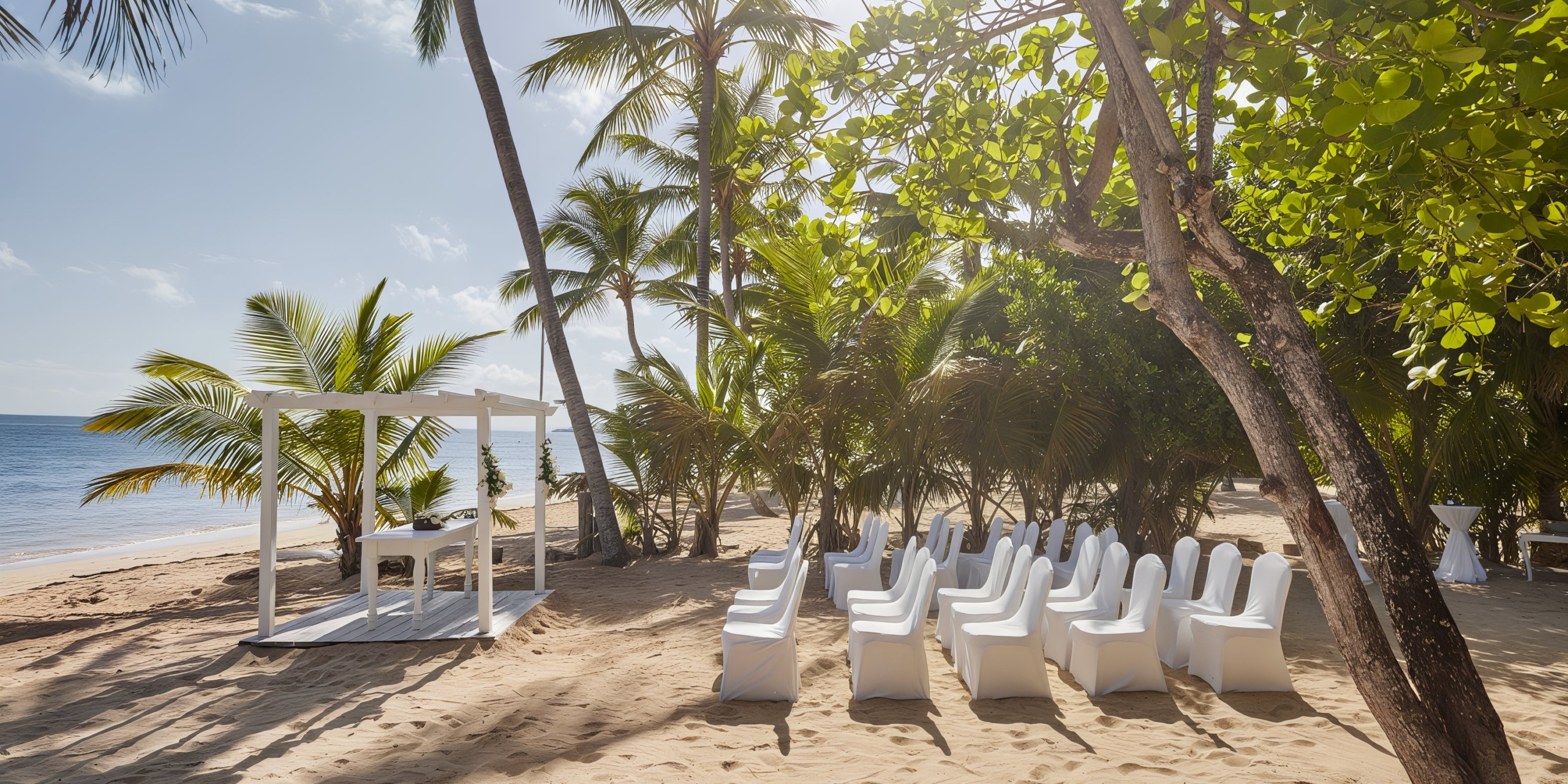 A beach wedding setup featuring white chairs arranged to face a charming small white gazebo, all surrounded by palm trees with a stunning ocean view at the Bahia Principe Grand Samana.