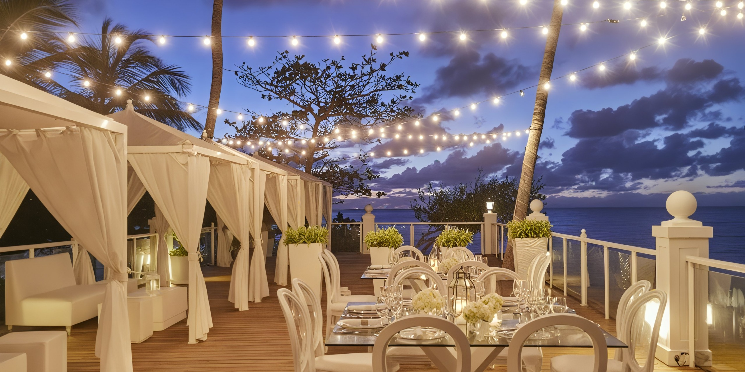 The outdoor dining area at Bahia Principe Grand Samana features white tables and chairs, covered cabanas, and string lights, all beautifully set against a twilight ocean backdrop adorned with palm trees.