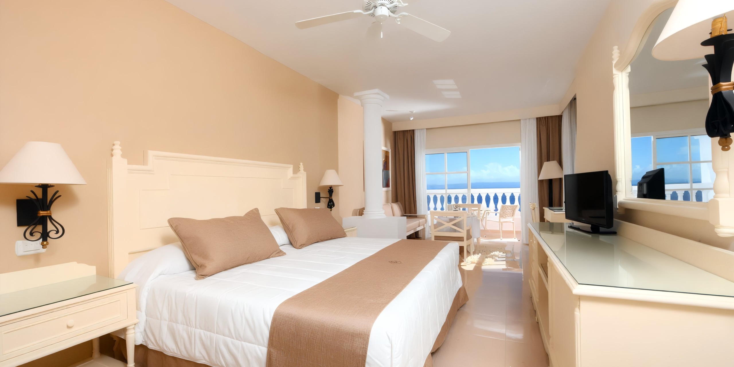 Experience the Bahia Principe Grand Samana's hotel room with a double bed adorned in beige accents, complete with a ceiling fan, a desk accompanied by a TV, and a balcony offering outdoor seating and captivating ocean views.
