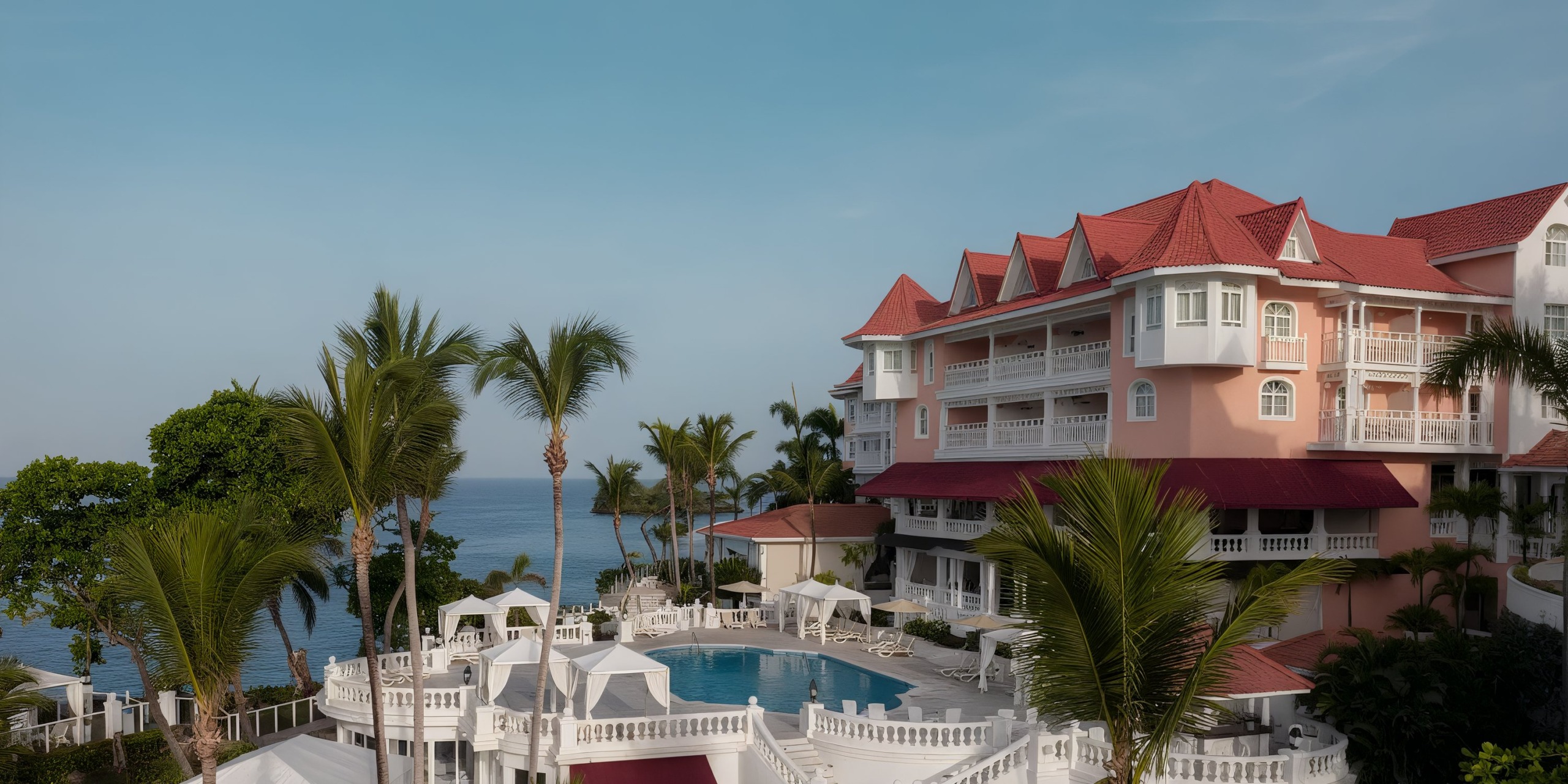 The Bahia Principe Grand Samana, featuring pink buildings with red roofs, overlooks a pool encircled by white cabanas and palm trees, all set against the stunning backdrop of the ocean and blue sky.