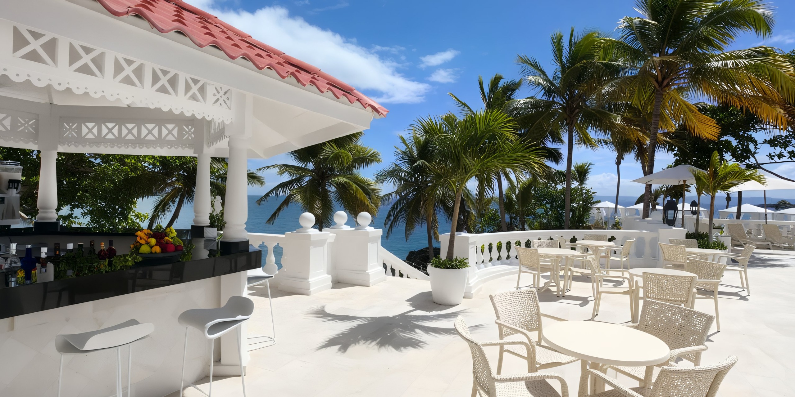 At Bahia Principe Grand Samana, you'll find an outdoor seaside bar featuring tables and chairs nestled under palm trees. The white architecture is enhanced with decorative details, set against a backdrop of the ocean and a clear blue sky.