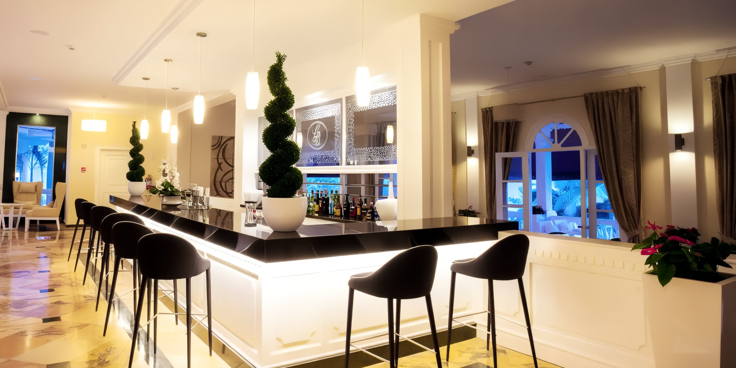 The Bahia Principe Grand Samana features a sleek, modern bar with black stools and an illuminated counter, enhanced by spiral topiary plants and a wide selection of bottles. The stylish setting is completed by large windows and elegant decor.