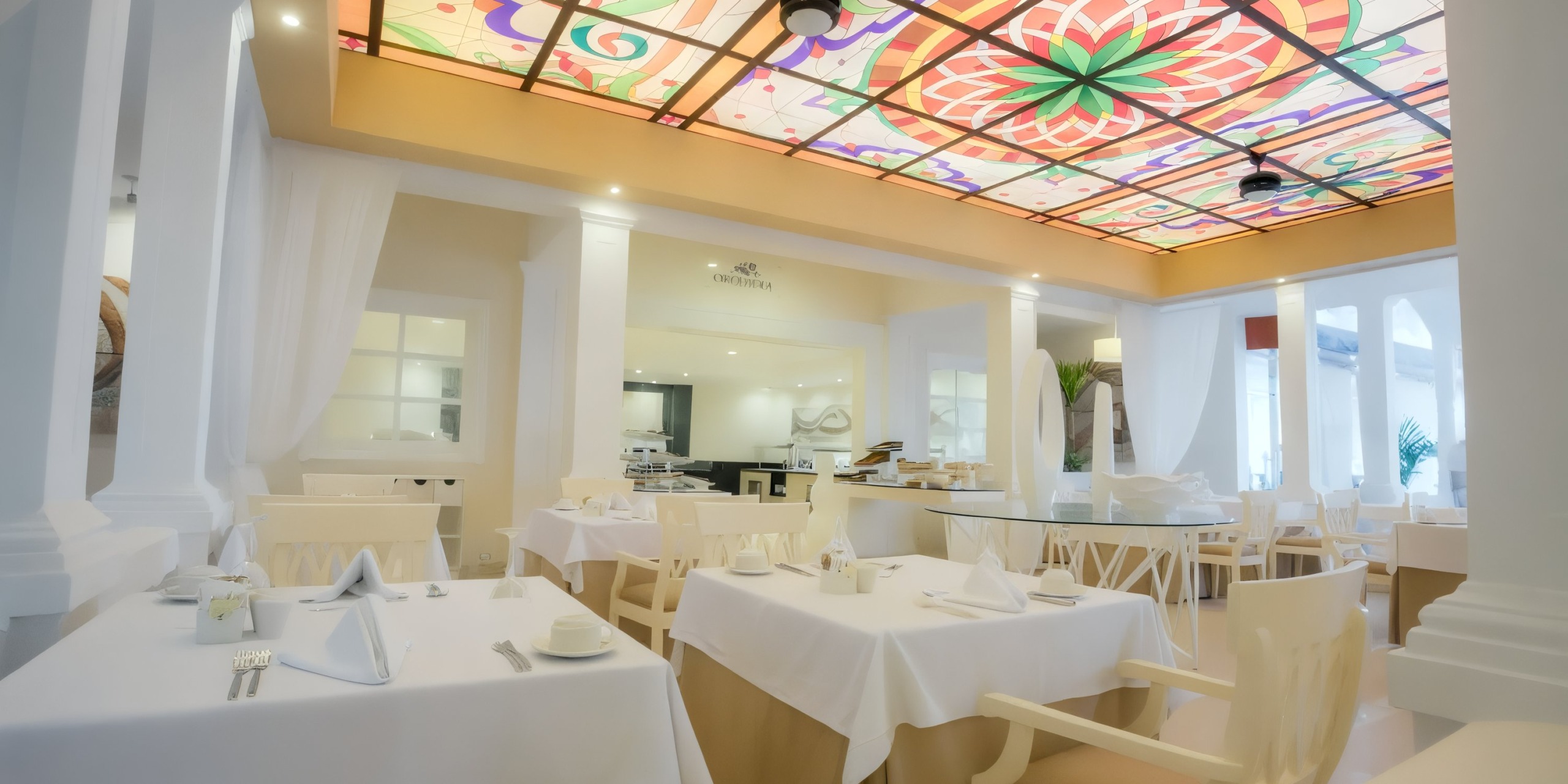 The interior of the Bahia Principe Grand Samana is bright and elegant, featuring white tablecloths on neatly arranged tables and a vibrant stained glass ceiling.