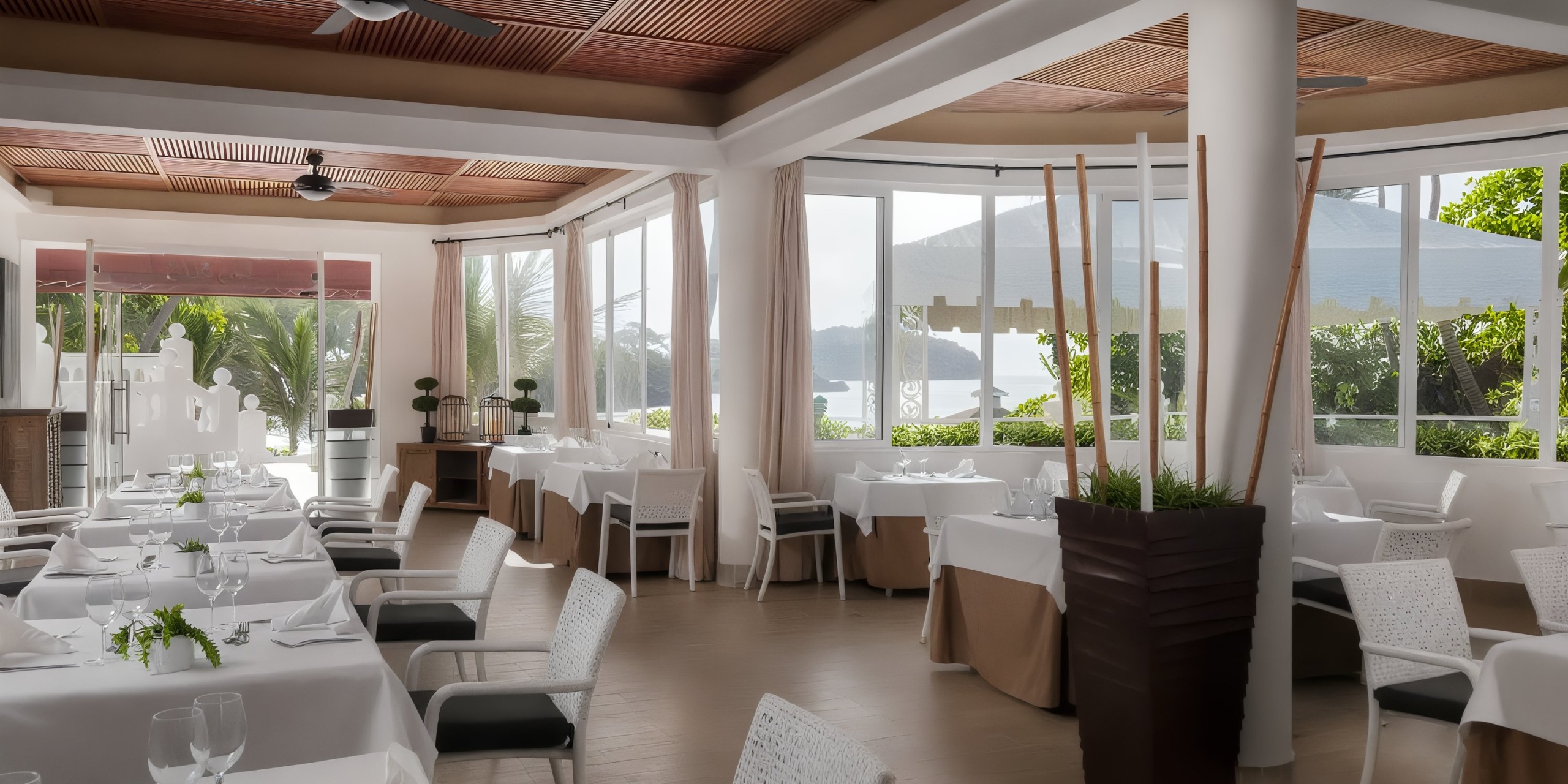 The interior of the Bahia Principe Grand Samana features an elegant restaurant setting with white tablecloths, modern decor, a wooden ceiling, and large windows that offer views of lush greenery and water outside.