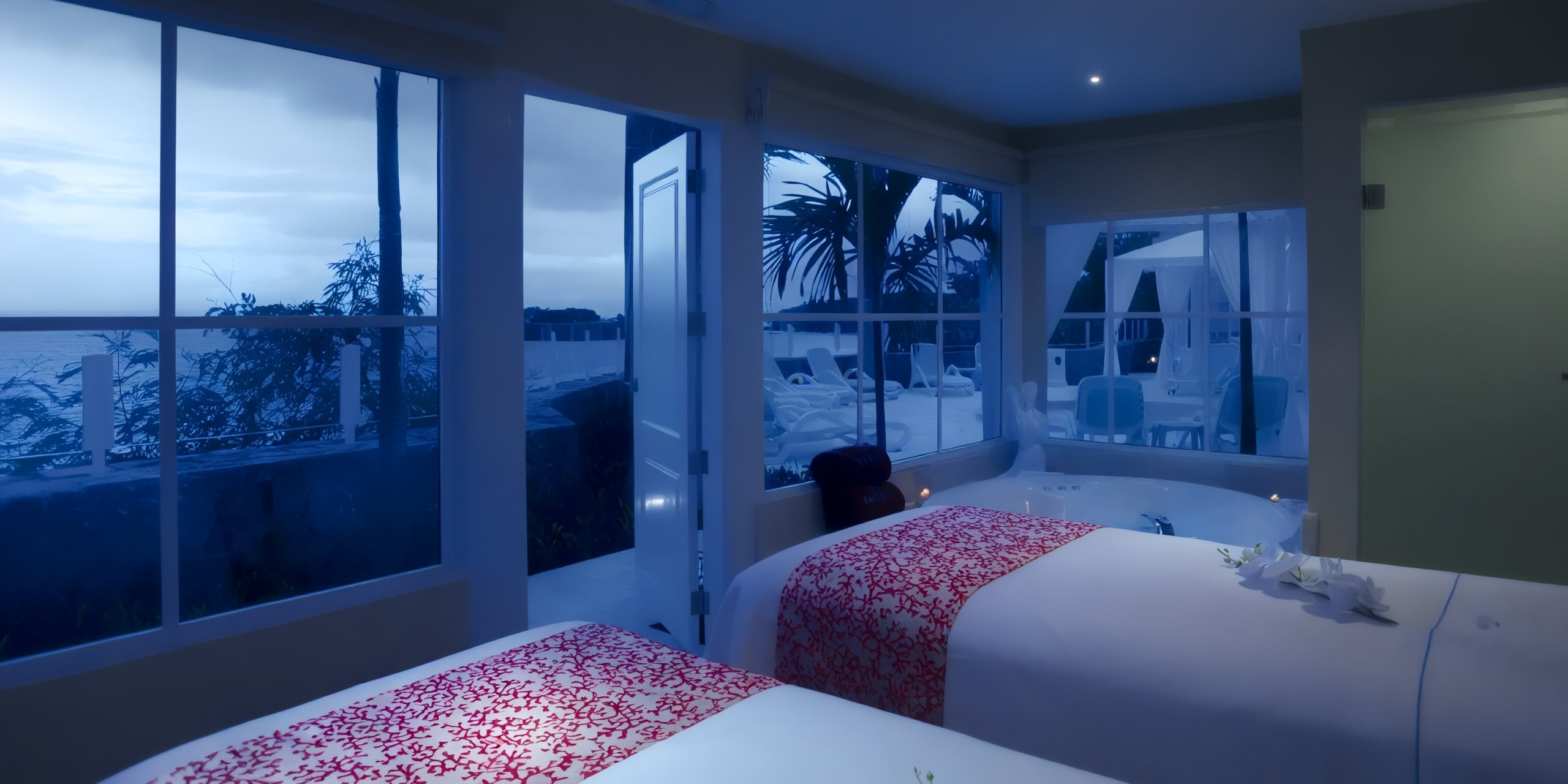 In the Bahia Principe Grand Samana, a tranquil spa room with two massage tables draped in red-patterned covers offers a view of the patio and ocean through expansive windows at twilight.