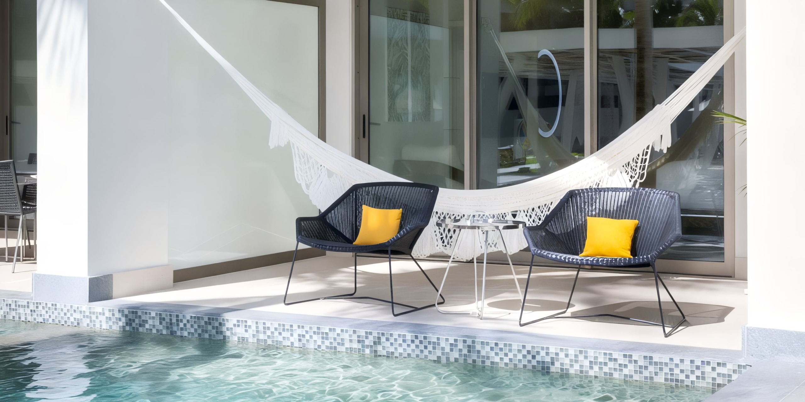 Garza Blanca Cancun's poolside patio features two elegant black chairs, each adorned with a vibrant yellow cushion, placed beside a small round table and accompanied by a pristine white hammock, all overlooking the clear blue waters of the swimming pool.