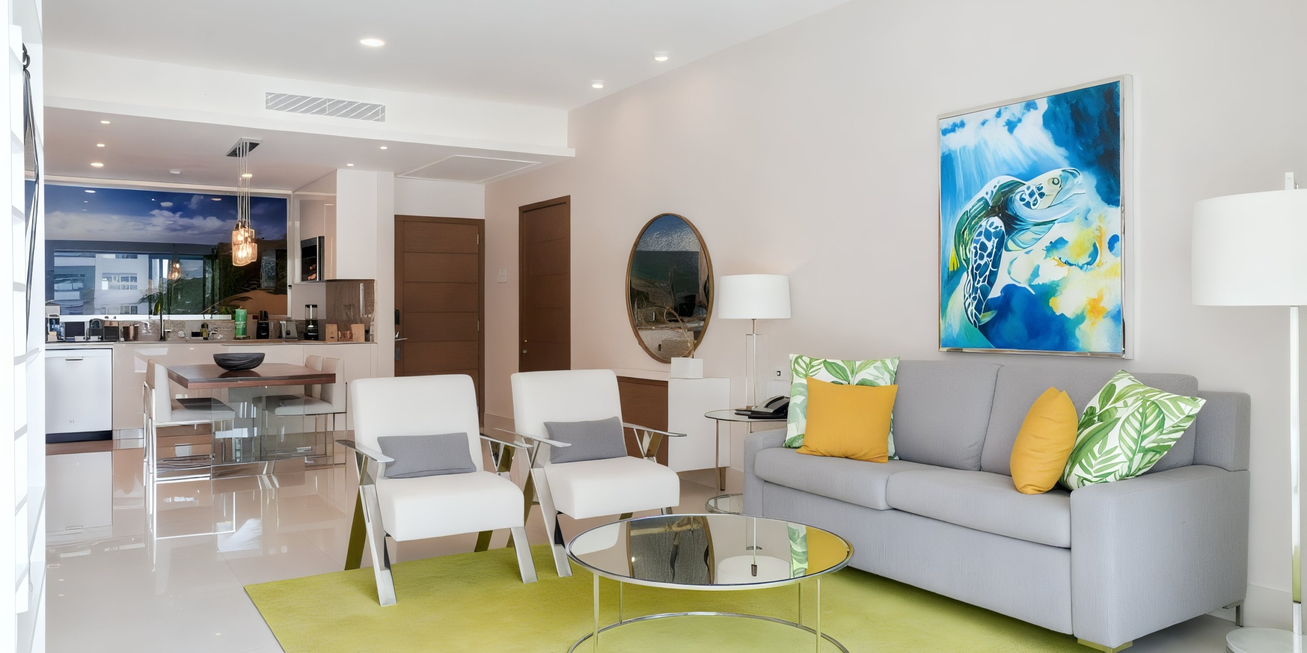The Garza Blanca Cancun features a contemporary living room complete with a gray sofa adorned with yellow and green pillows, a sleek glass coffee table, two stylish chairs, and an impressive large painting of a turtle. In the background, you'll find an open kitchen with a dining table.