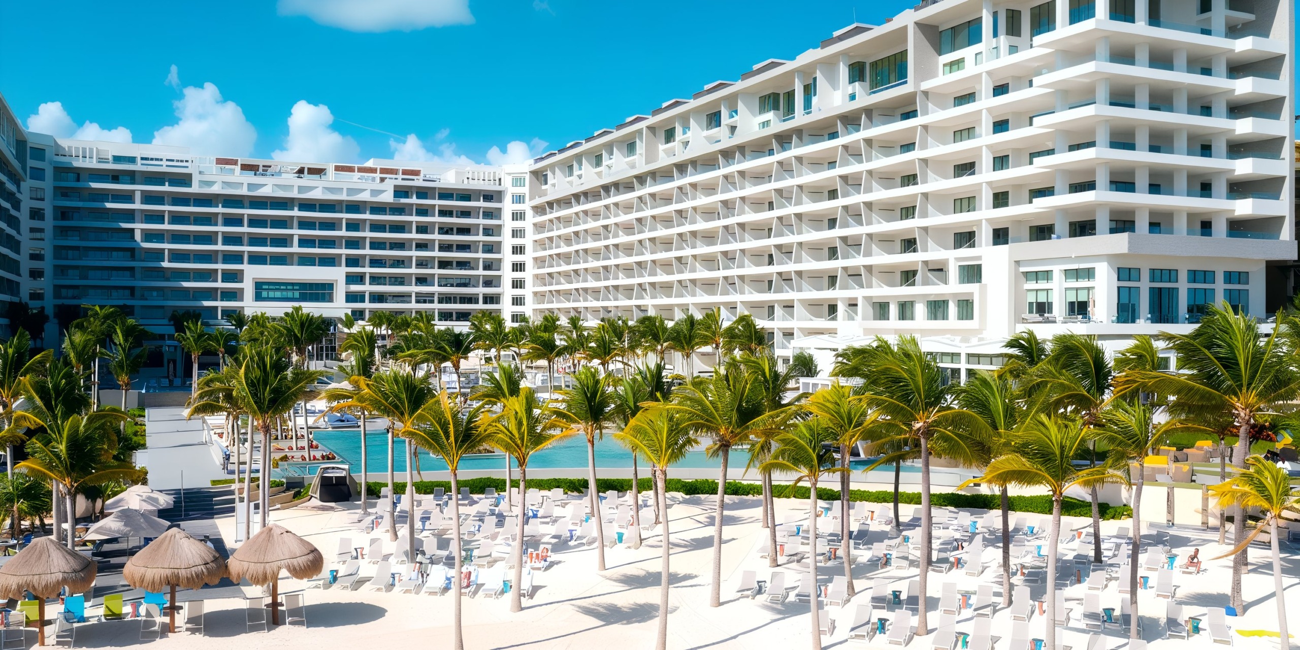 Garza Blanca Cancun, a large beachfront hotel with a white exterior and surrounded by palm trees, overlooks a spacious pool area equipped with lounge chairs and umbrellas beneath a clear blue sky.