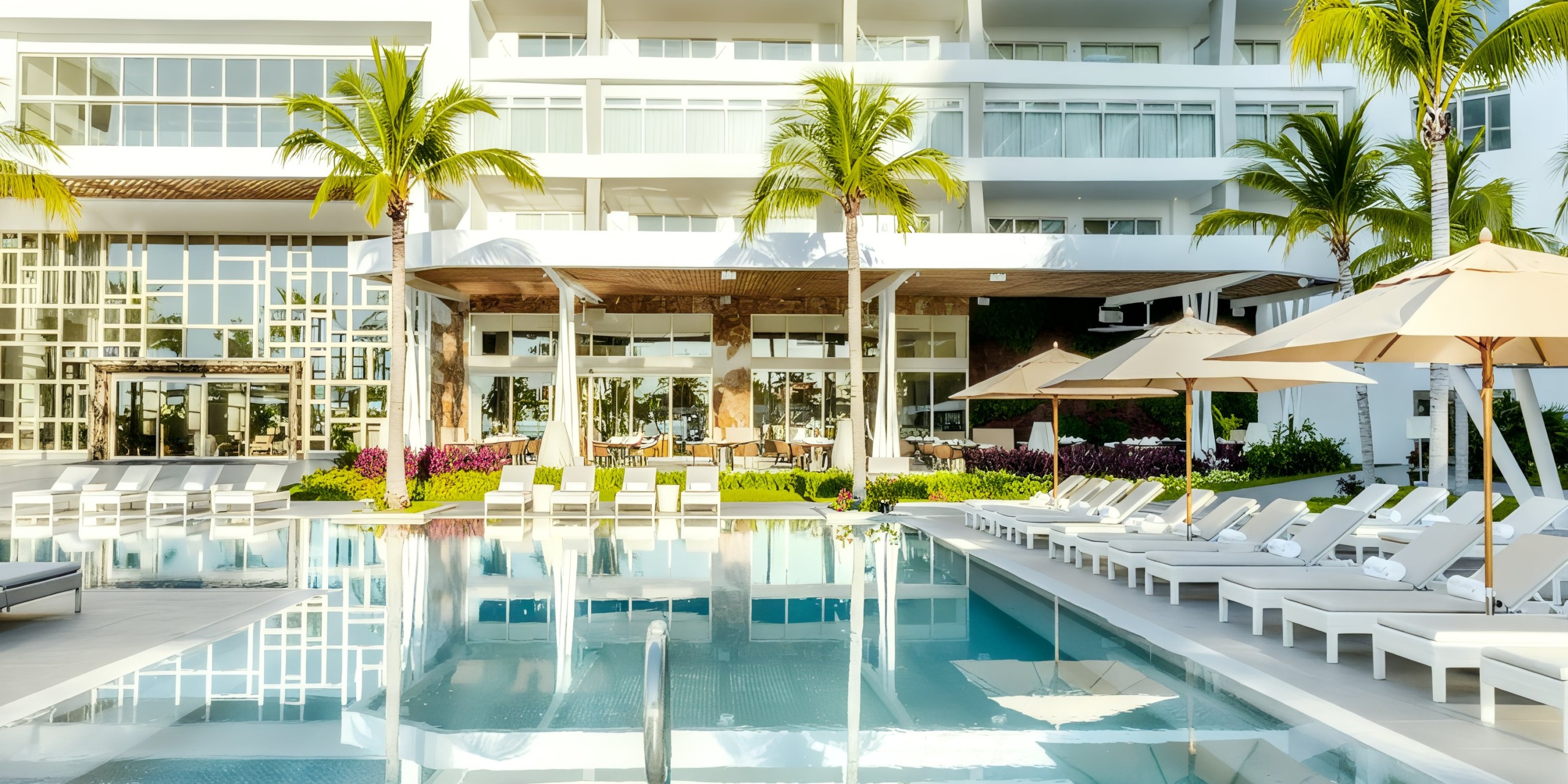 Experience the luxurious pool area at Garza Blanca Cancun, equipped with sun loungers, umbrellas, and palm trees, all set against a backdrop of the resort's white facade and glass windows.