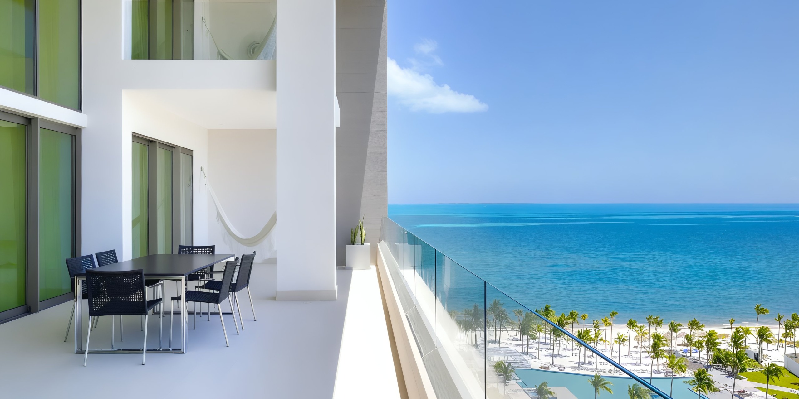 At Garza Blanca Cancun, a contemporary balcony featuring a glass railing provides stunning views of the beach and ocean. It is equipped with a dining table, chairs, a hammock, and is complemented by palm trees lining the shore.