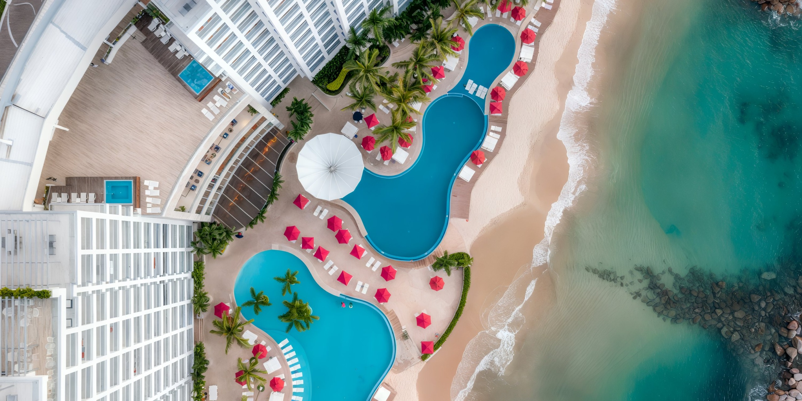 Aerial view of the Hilton Vallarta Riviera All-Inclusive Resort featuring curvy swimming pools surrounded by red umbrellas and lounge chairs, situated next to a sandy beach and ocean.