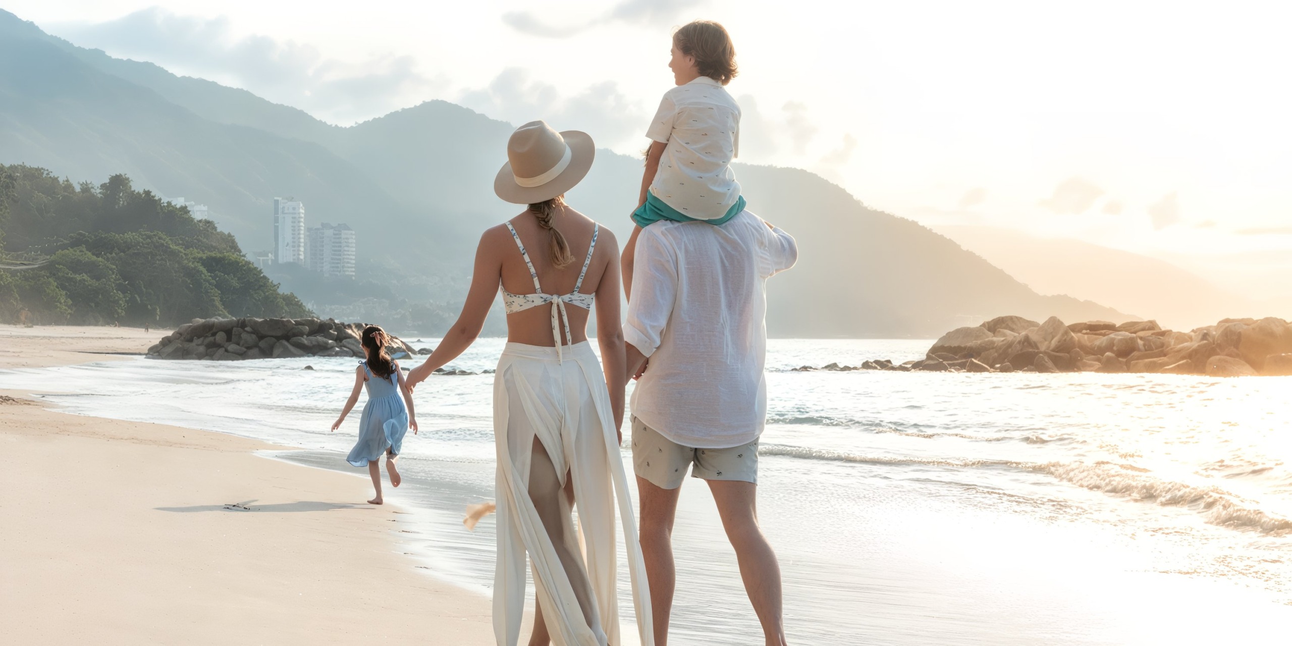A family of four strolls along the sandy beach at the Hilton Vallarta Riviera All-Inclusive Resort, with mountains and buildings visible in the distance. One child enjoys a ride on a man's shoulders while another runs happily ahead along the shore.
