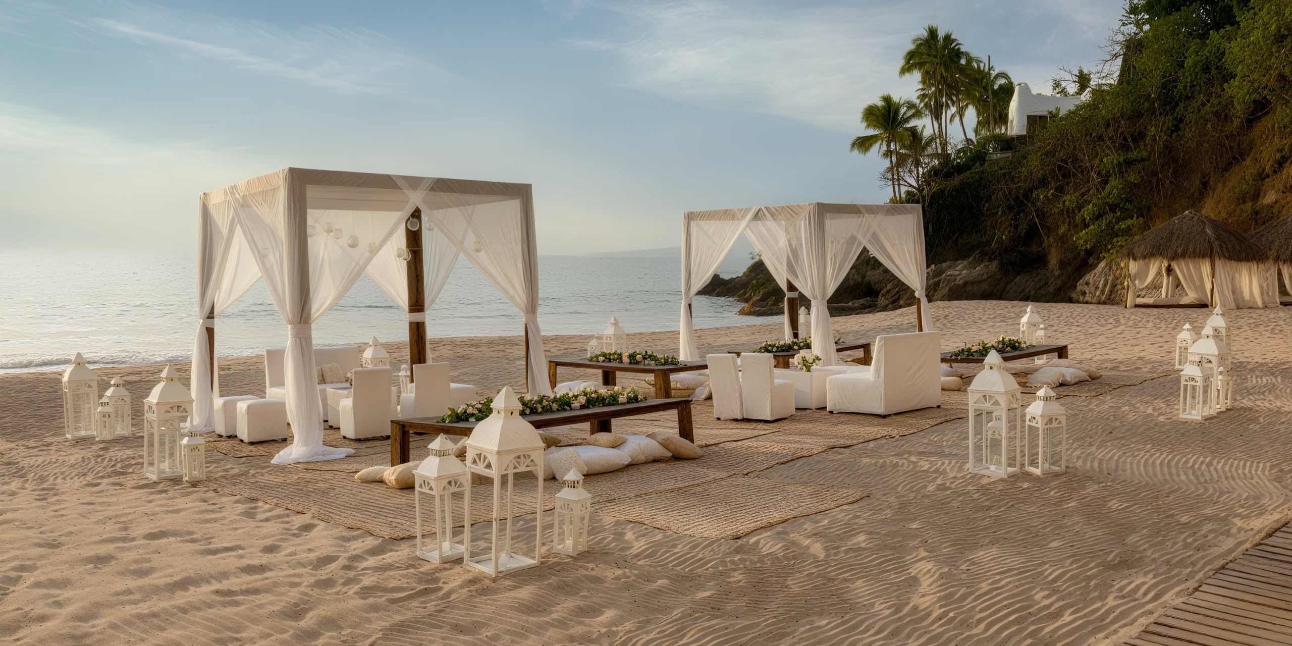 Relax in style at the Hilton Vallarta Riviera All-Inclusive Resort with an elegant beach setup featuring two canopied seating areas, chic white furniture, and charming lanterns on the sandy shore by the ocean; all framed by picturesque palm trees.