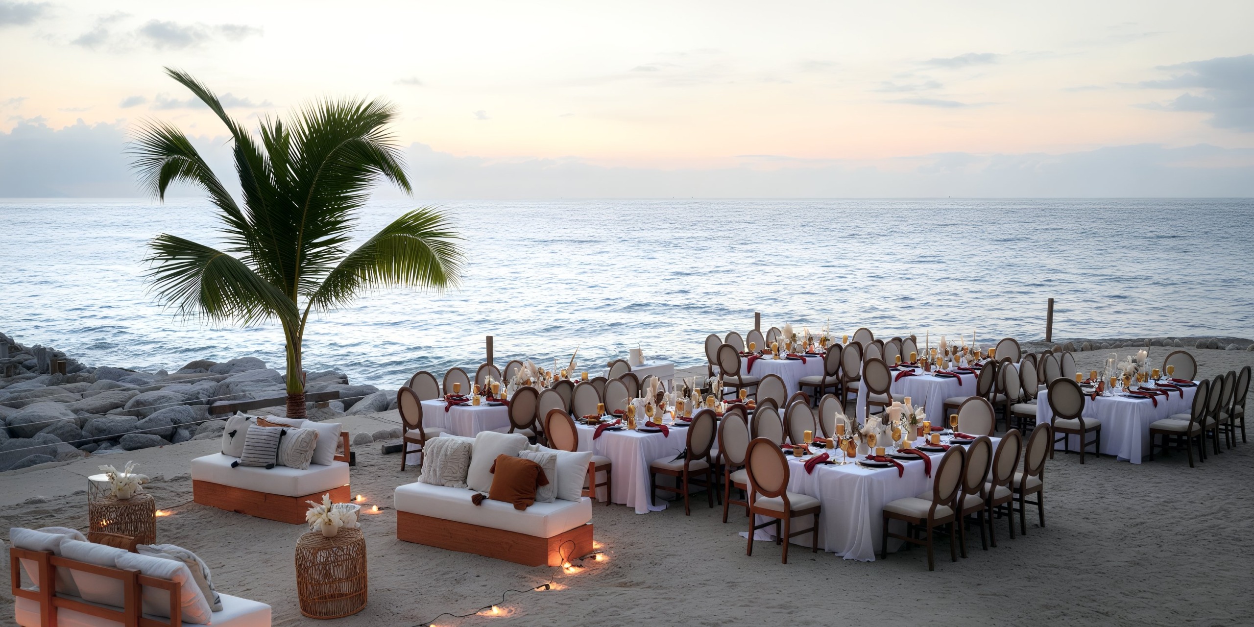 Experience an enchanting outdoor beachside dining setup at the Hilton Vallarta Riviera All-Inclusive Resort, featuring round tables, chairs, and elegant decorative table settings on a sandy shore. Enjoy the serene backdrop of a palm tree and the ocean during sunset.