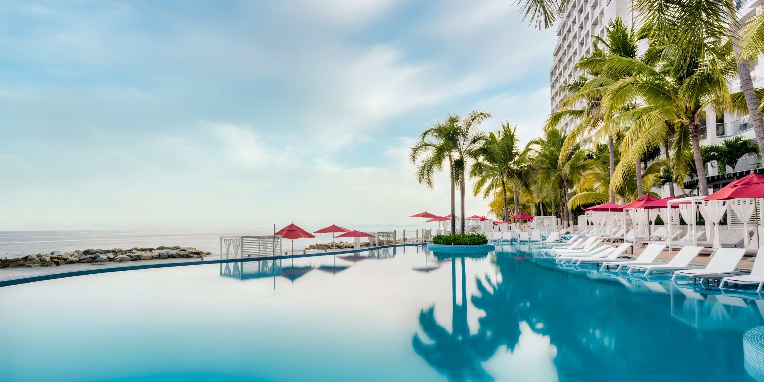 At the Hilton Vallarta Riviera All-Inclusive Resort, you can enjoy a tranquil poolside setting with palm trees and lounge chairs beneath red umbrellas, all with a stunning ocean view on a clear day.