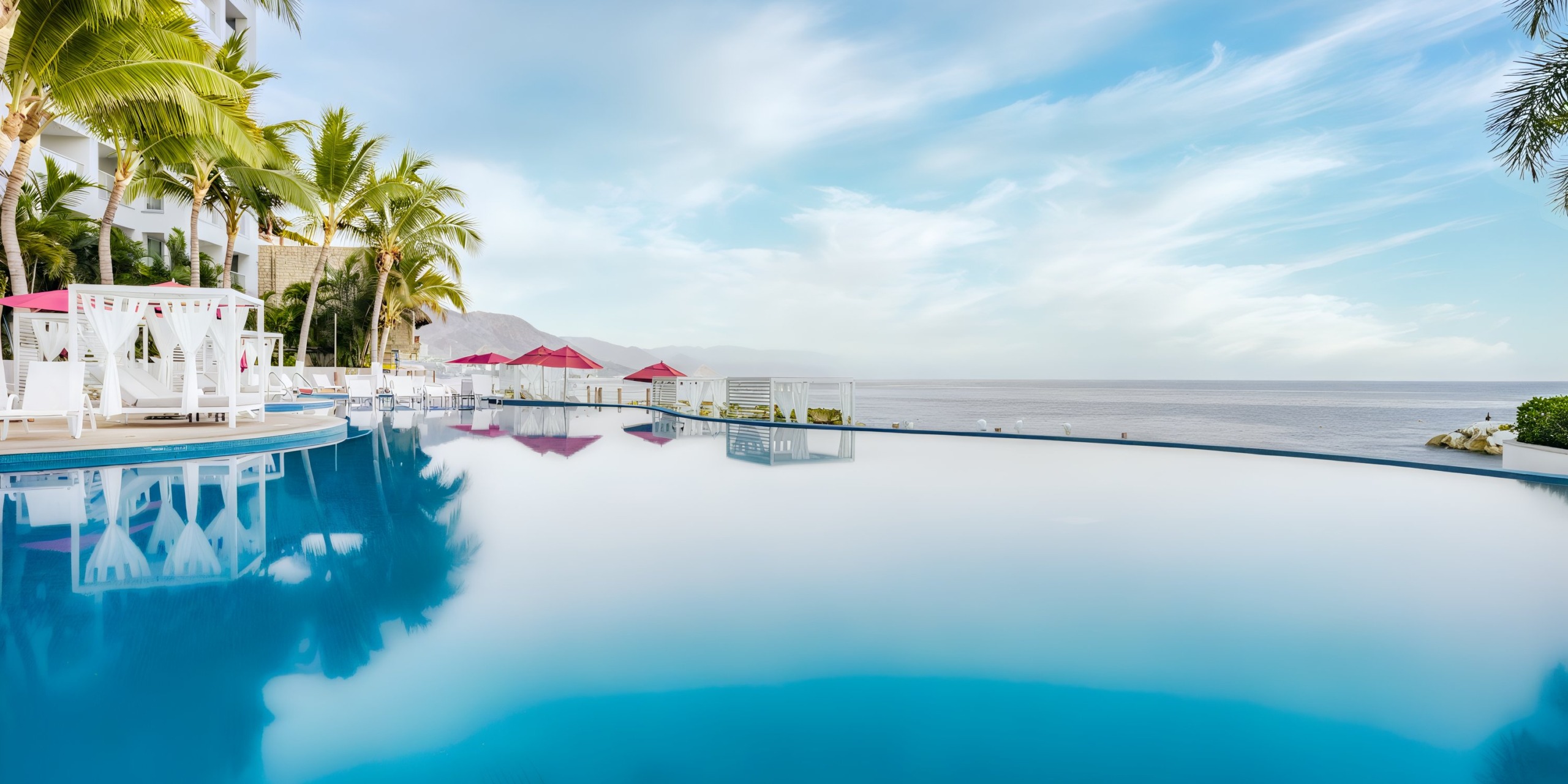 At the Hilton Vallarta Riviera All-Inclusive Resort, the infinity pool features clear blue water surrounded by white lounge chairs and palm trees, offering a stunning view of the ocean and distant mountains under a partly cloudy sky.