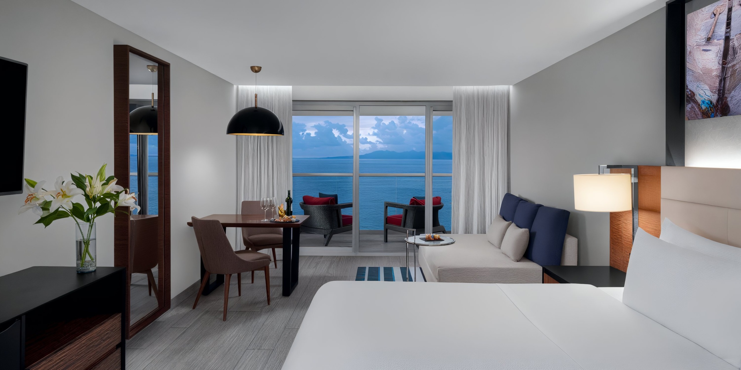 Enjoy a modern room at the Hilton Vallarta Riviera All-Inclusive Resort, complete with a comfortable bed, seating area, and dining table. A sliding glass door opens up to a balcony with stunning views of the ocean.