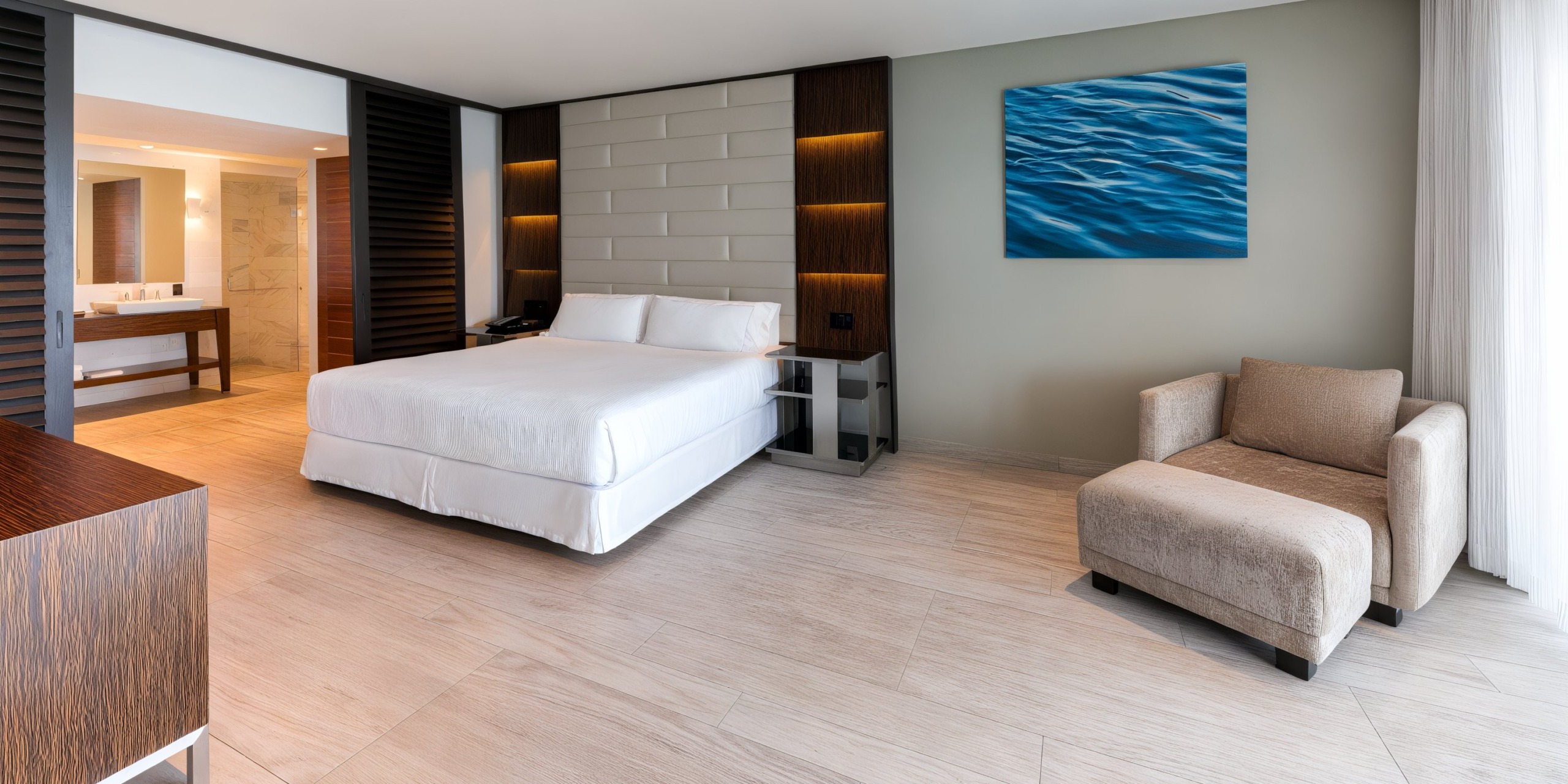 Experience a contemporary room at the Hilton Vallarta Riviera All-Inclusive Resort, featuring a spacious bed, cozy armchair, and wall art depicting a serene water scene. Enjoy the elegant wooden flooring and an open bathroom area complete with a vanity and mirror.