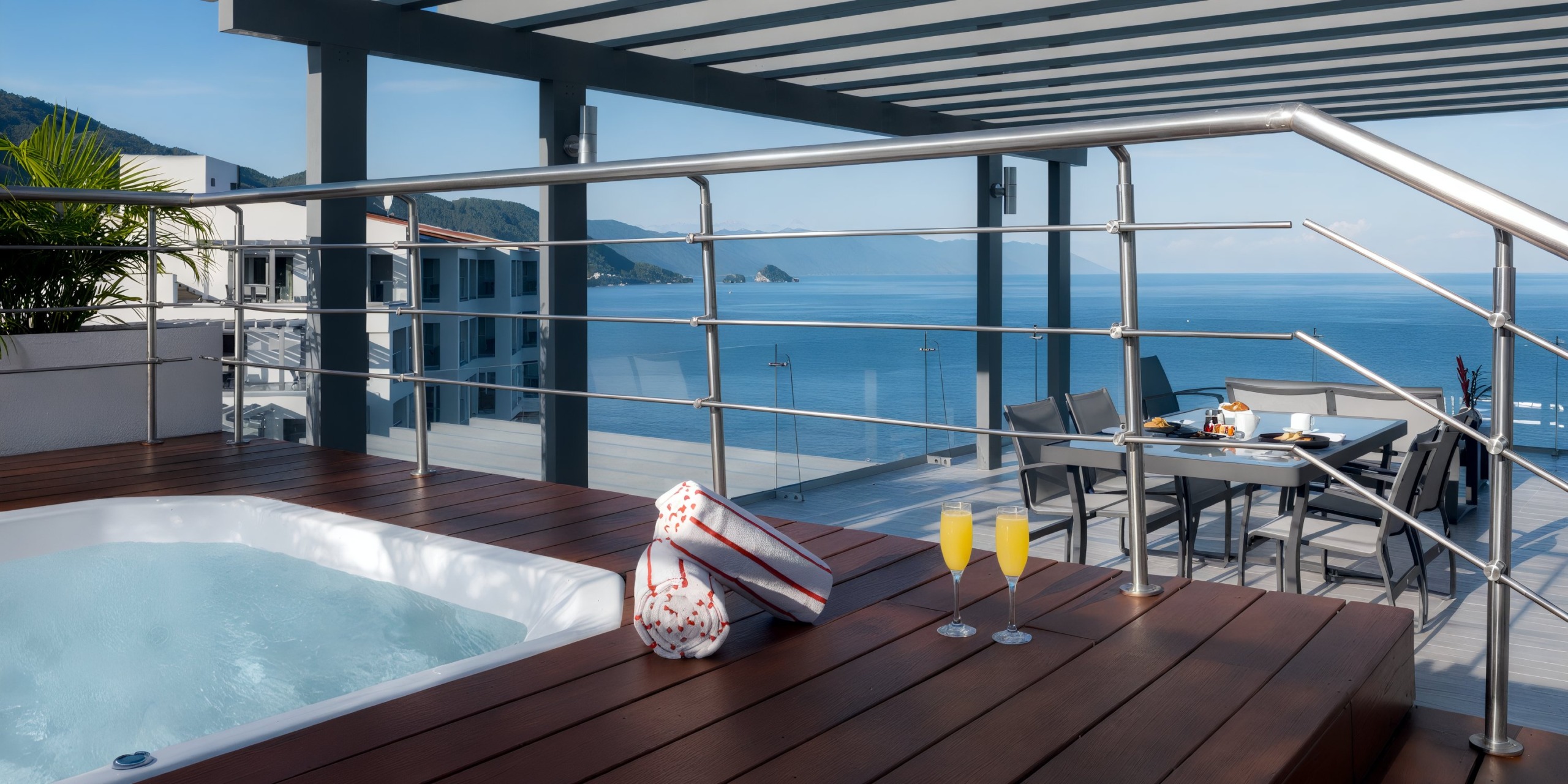 Enjoy a rooftop deck at Hilton Vallarta Riviera All-Inclusive Resort featuring a hot tub, a table set with breakfast items, and two mimosas, all while overlooking the ocean and distant mountains.